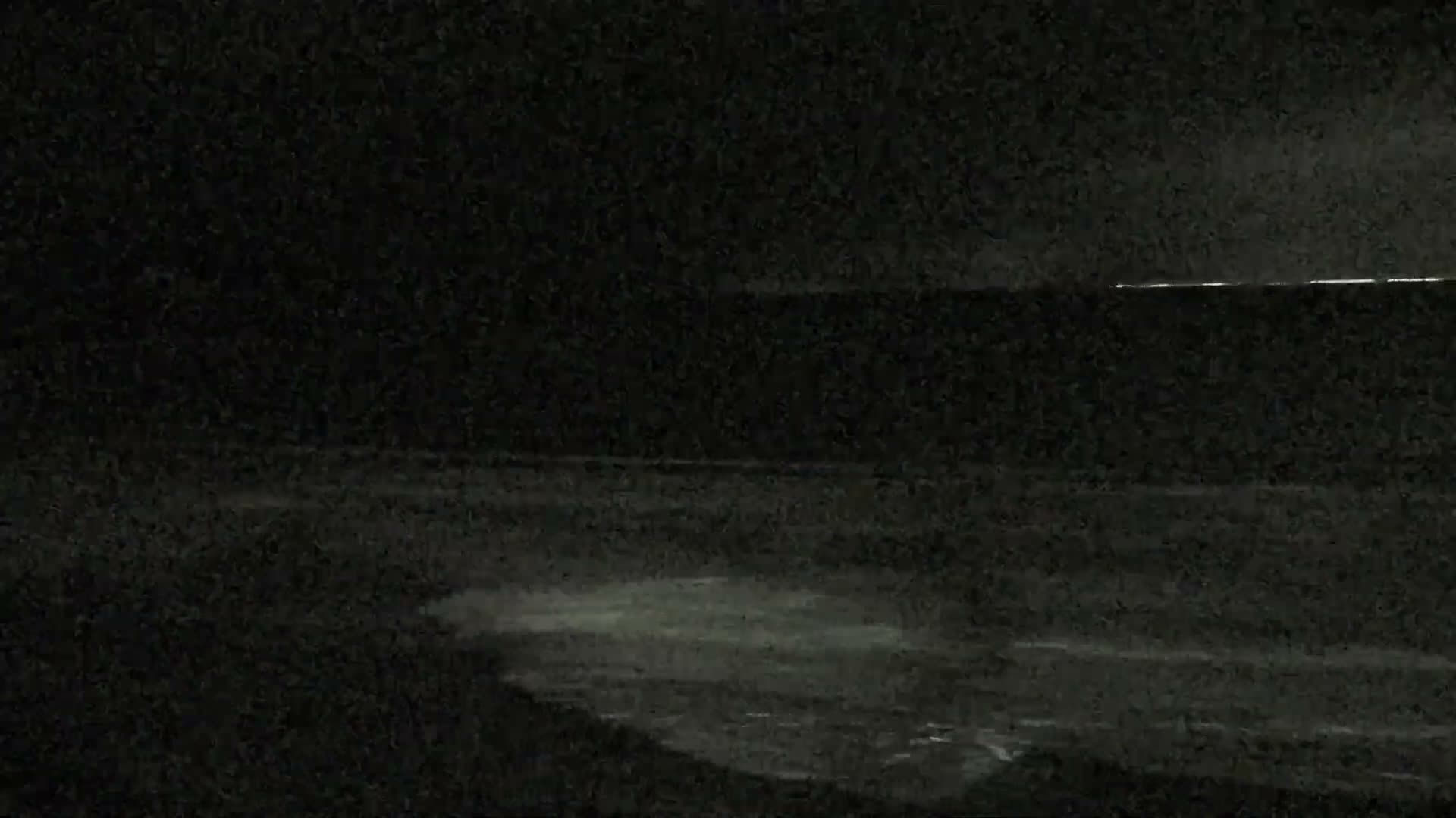 A coastal road with parked cars and benches runs alongside a grassy slope leading to a sandy bay with gentle waves, all under a partly cloudy sky, with houses and dense trees visible along the distant shoreline.