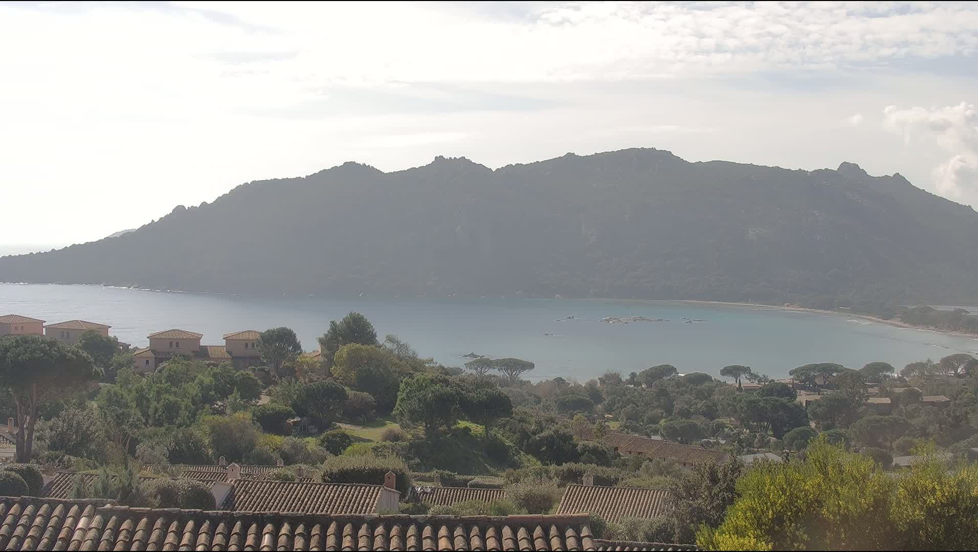 A hazy coastal scene shows a calm bay surrounded by a mountain range and dotted with houses, all under a mostly clear sky.