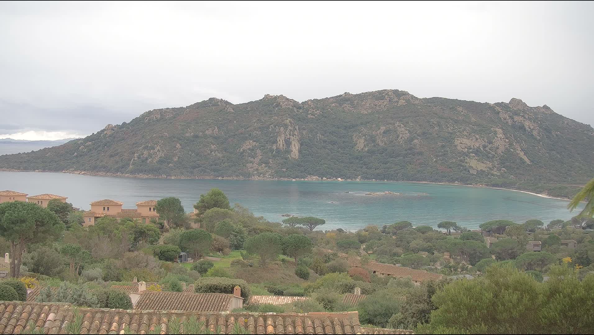 Under an overcast sky, a scenic coastal landscape reveals a turquoise bay bordered by a sandy beach and a rugged, tree-covered mountain range, with numerous terracotta-roofed homes nestled among lush green foliage in the foreground.