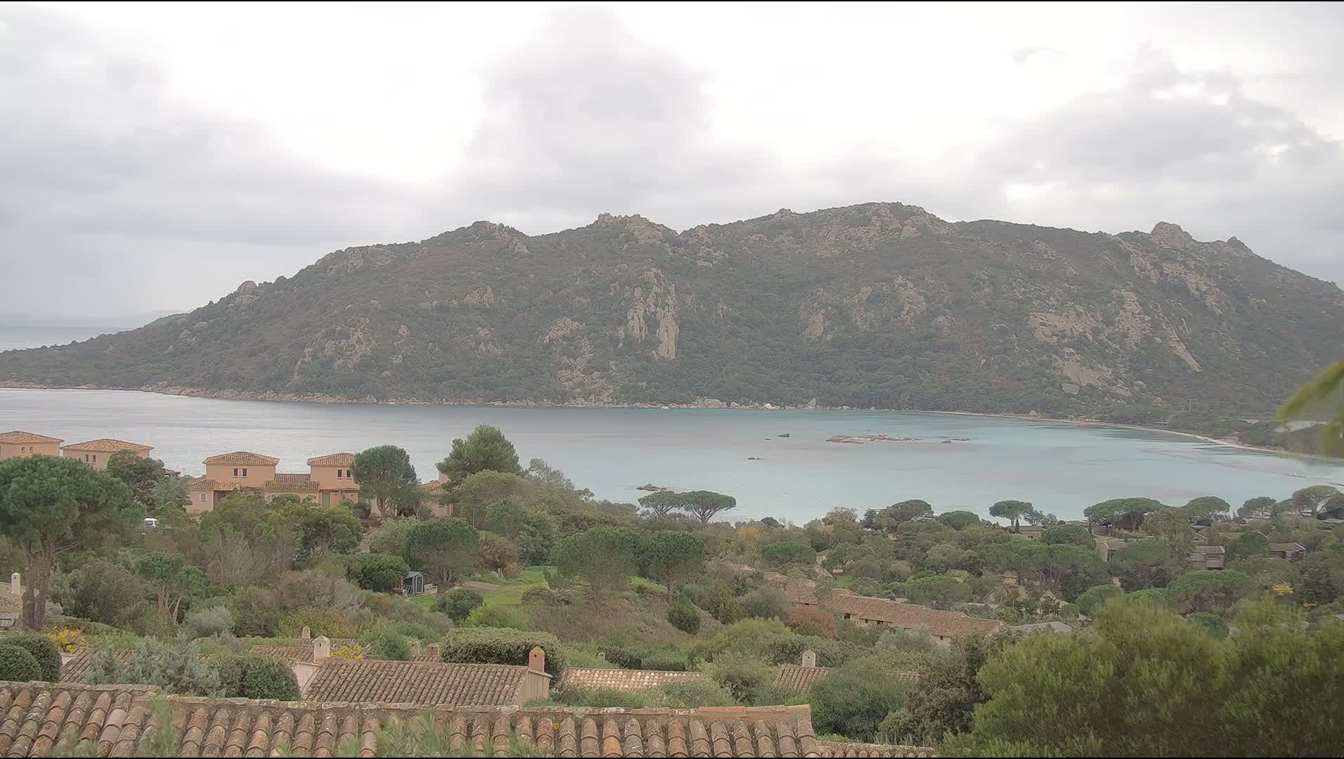 Under an overcast sky, a wide bay with calm, light blue-green water is nestled before a large, rocky, tree-covered mountain range, while the foreground displays terracotta rooftops, light-colored buildings, and dense greenery.