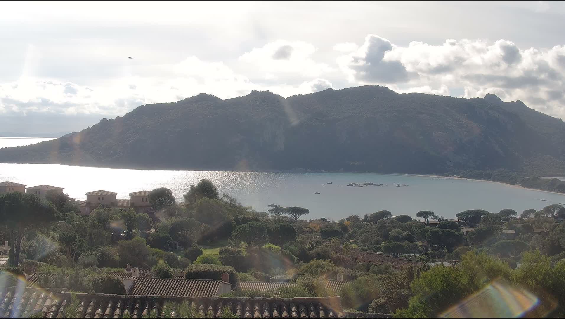 Under an overcast sky, a wide bay with calm, light blue-green water is nestled before a large, rocky, tree-covered mountain range, while the foreground displays terracotta rooftops, light-colored buildings, and dense greenery.
