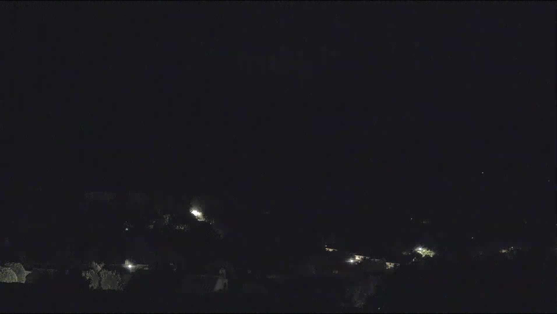 Under an overcast sky, a wide bay with calm, light blue-green water is nestled before a large, rocky, tree-covered mountain range, while the foreground displays terracotta rooftops, light-colored buildings, and dense greenery.