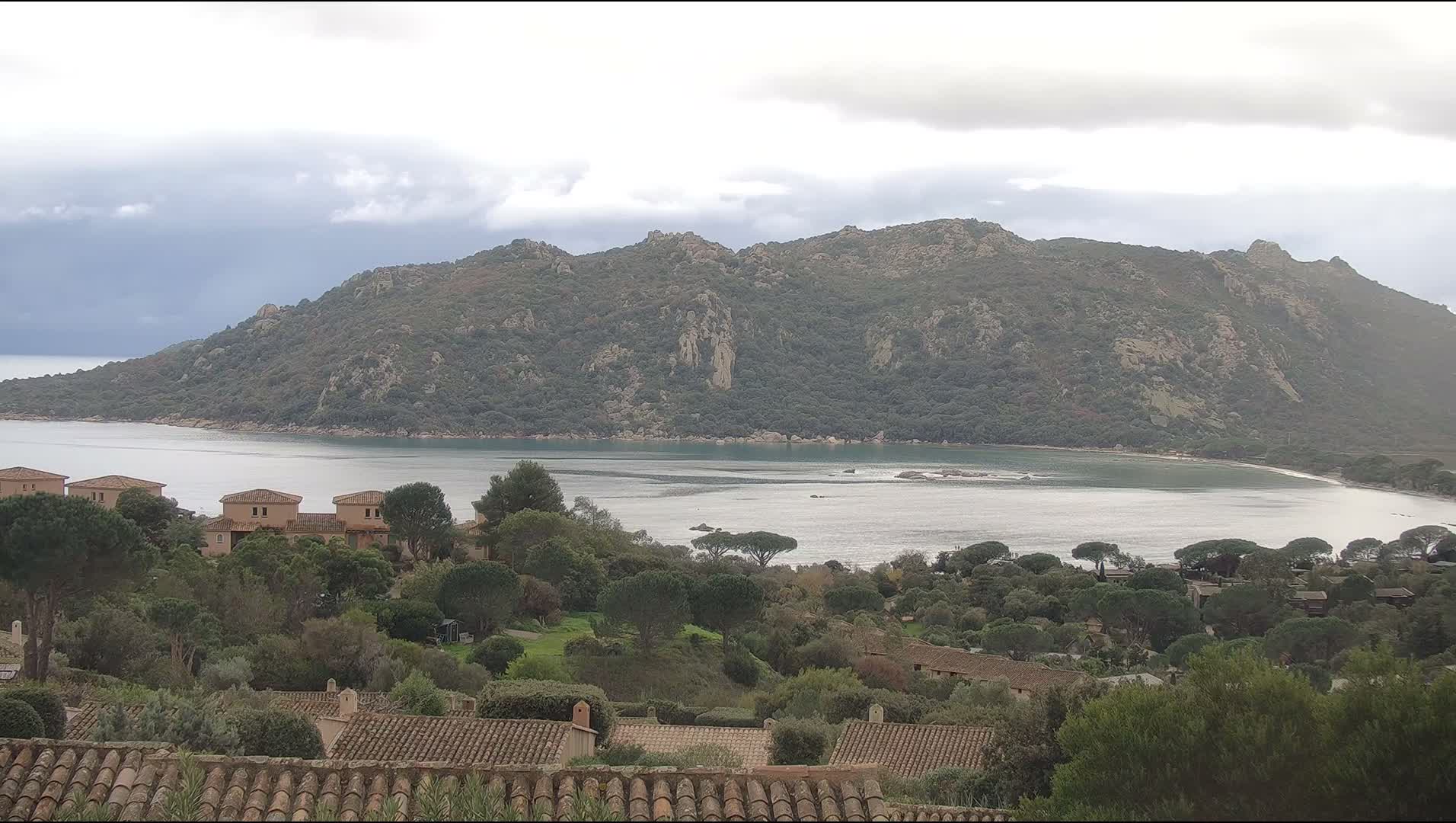 Under an overcast sky, a wide bay with calm, light blue-green water is nestled before a large, rocky, tree-covered mountain range, while the foreground displays terracotta rooftops, light-colored buildings, and dense greenery.