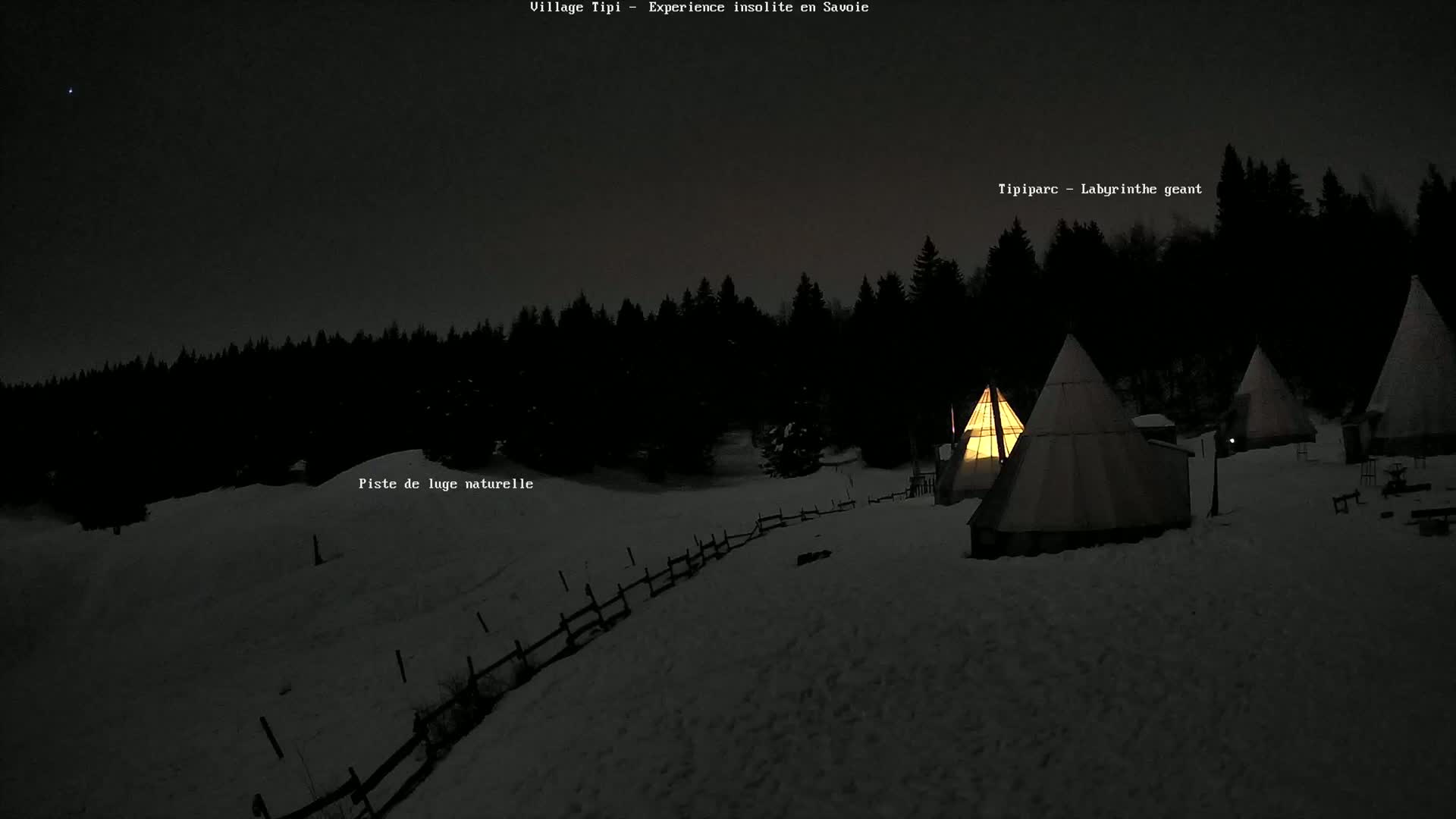 Several illuminated teepees are situated on a snow-covered landscape at night, backed by a dark forest under a clear sky.
