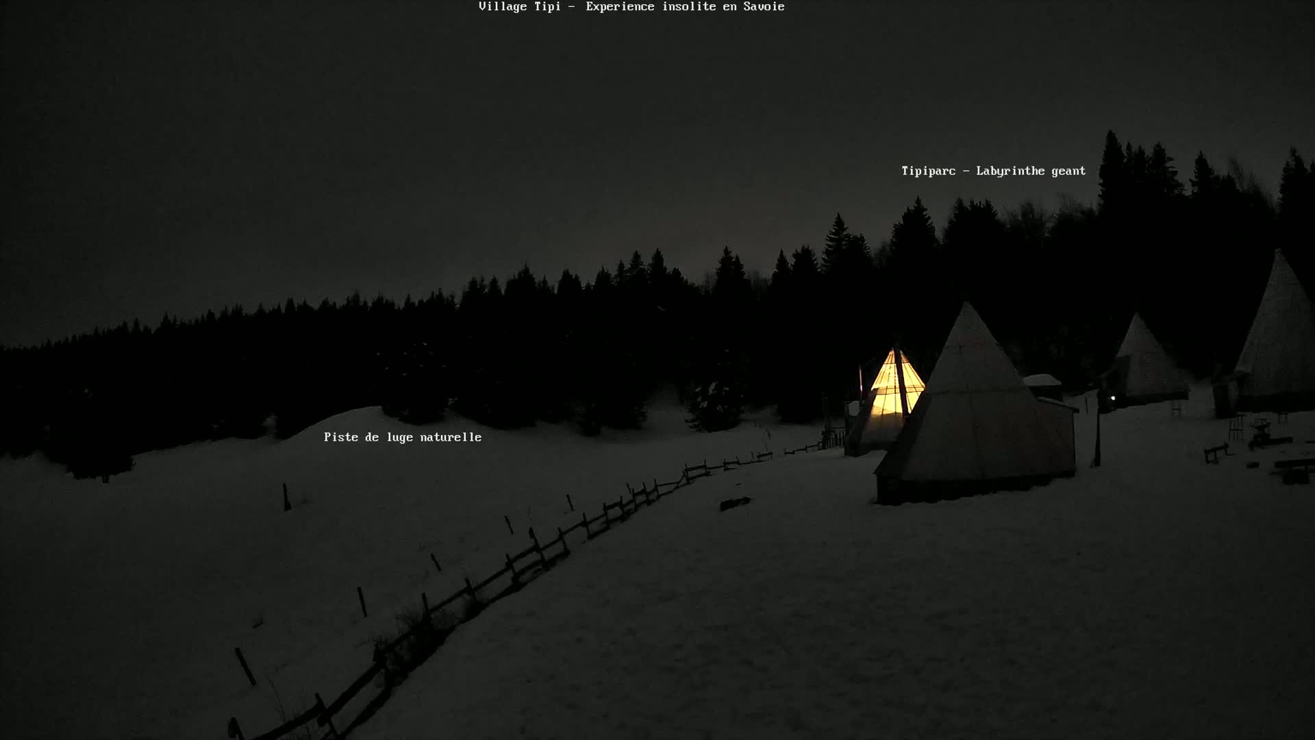Illuminated teepees are nestled in a snow-covered landscape at night, surrounded by a dark forest.