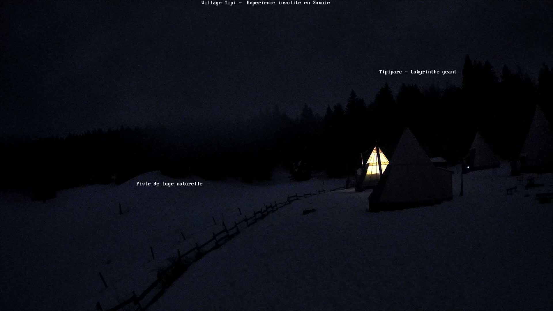 Illuminated teepees are nestled in a snow-covered landscape at night, surrounded by a dark forest under a dusky sky.