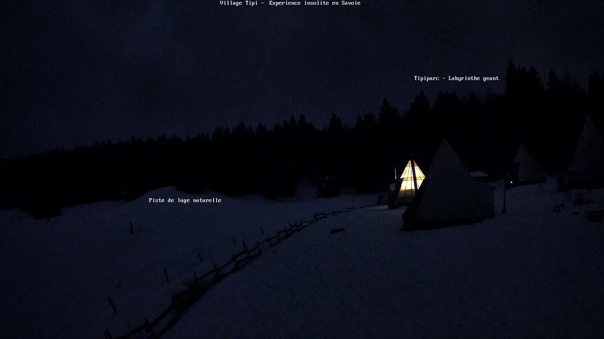 Illuminated teepees sit on a snow-covered landscape at night, surrounded by a dark forest.