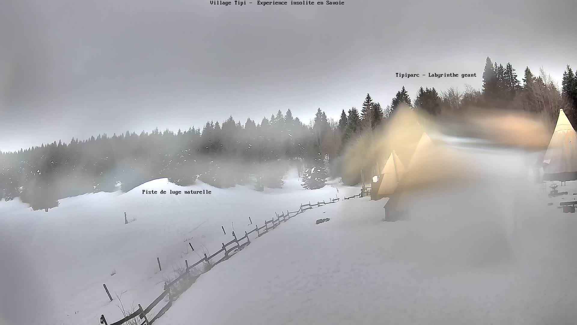 A snowy, foggy landscape features several teepee-like structures behind a wooden fence, with a forest in the background.