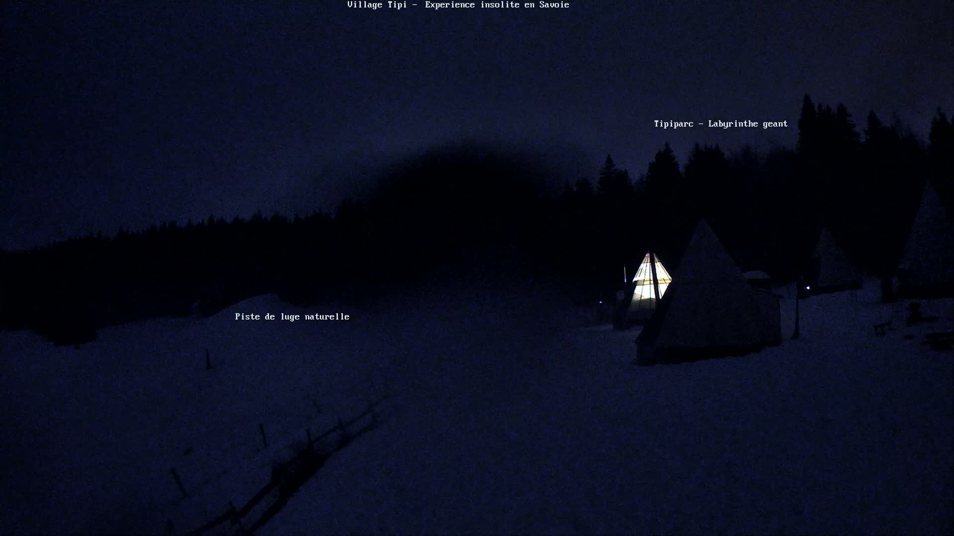 A snow-covered landscape at night, illuminated by the warm light of several teepees nestled amongst dark trees.