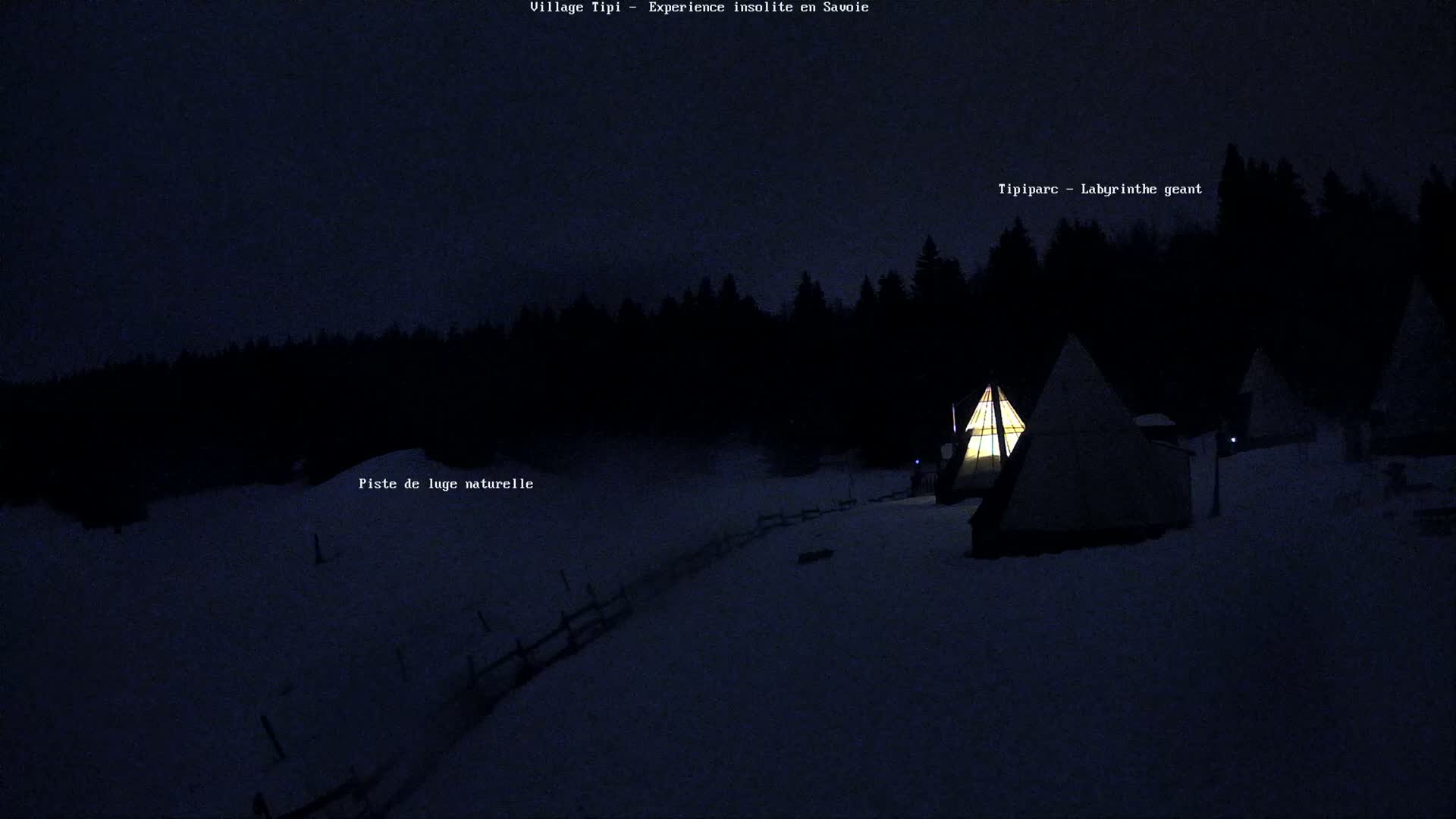 A snow-covered landscape at night, illuminated by the warm glow of several lit teepees, is bordered by a dark forest.
