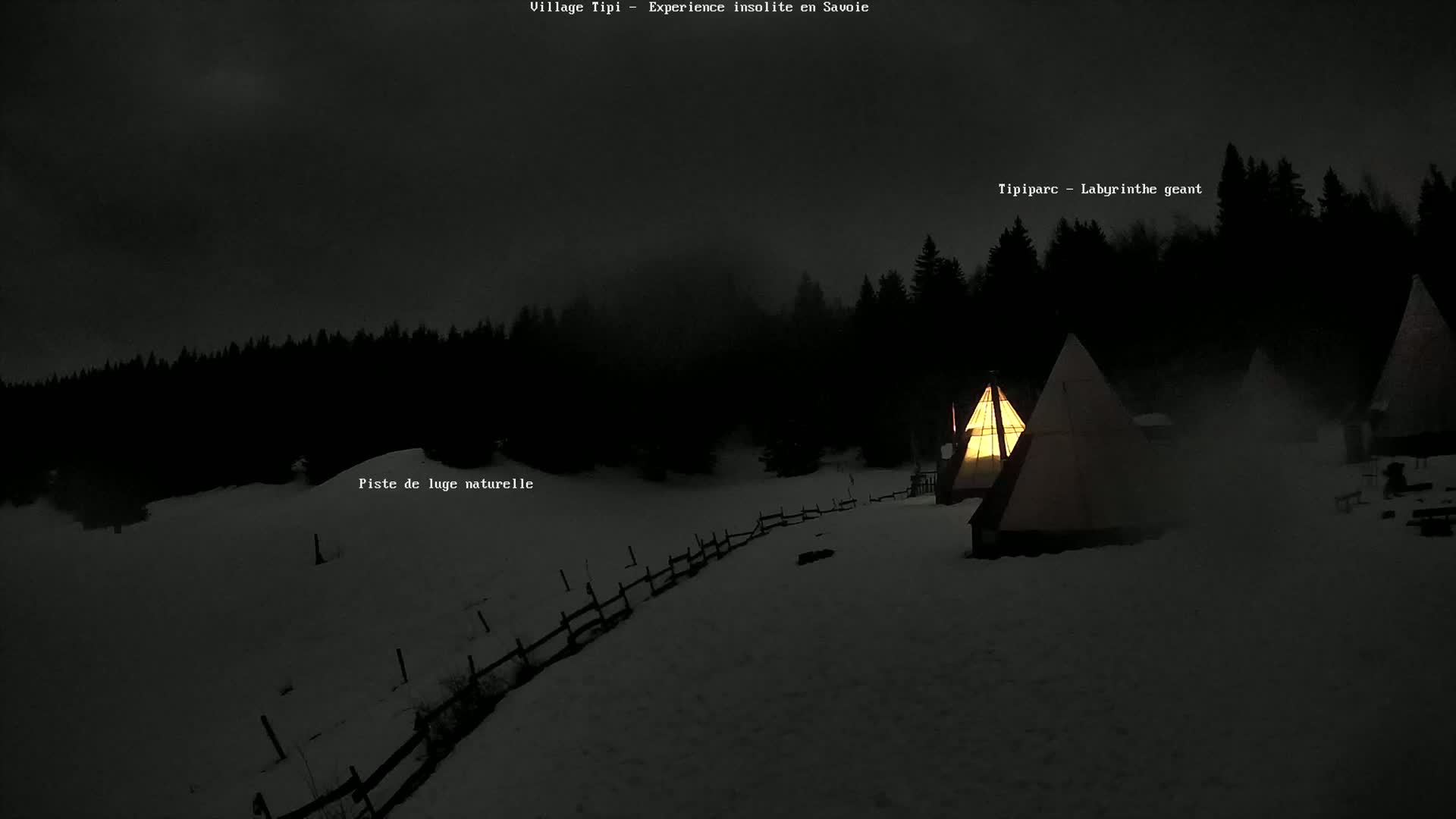 Illuminated teepees stand on a snow-covered landscape at night, surrounded by a dark forest under a mostly clear sky.