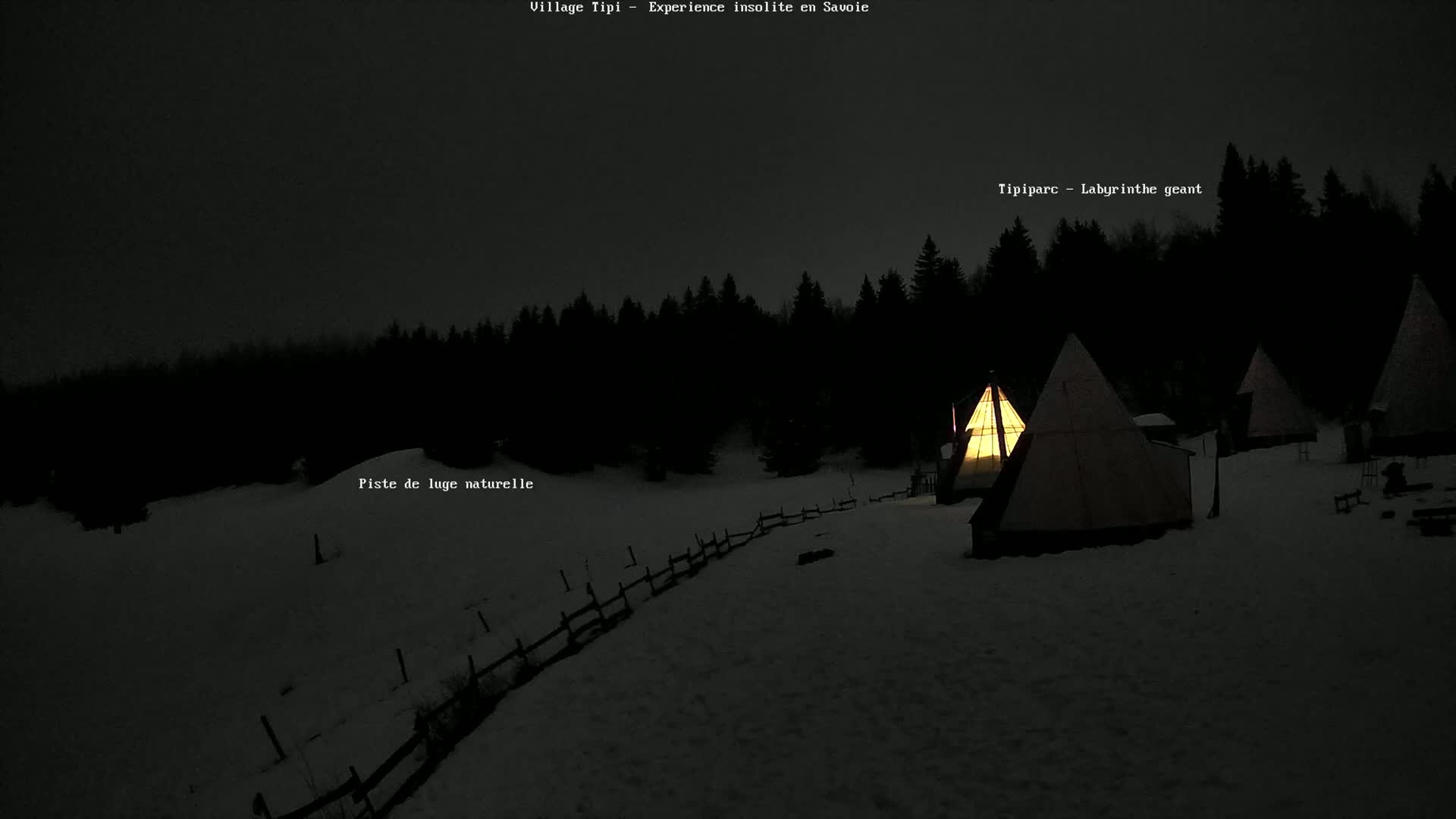 Several illuminated teepees sit on a snow-covered landscape at night, surrounded by a dark forest under a dark sky.