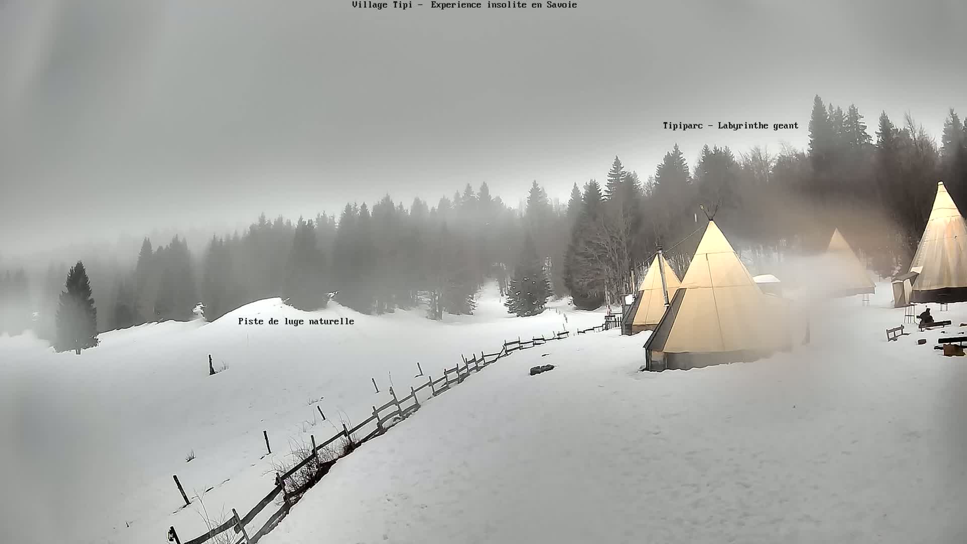 Several teepees sit in a snowy landscape shrouded in fog, with a wooden fence visible in the foreground.