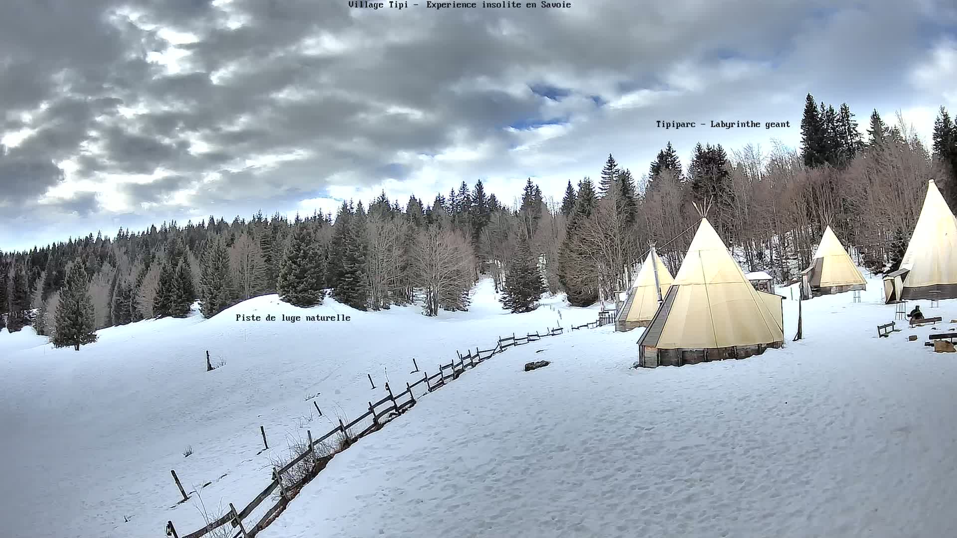 Several teepees are situated on a snow-covered landscape in front of a coniferous forest under a cloudy sky.
