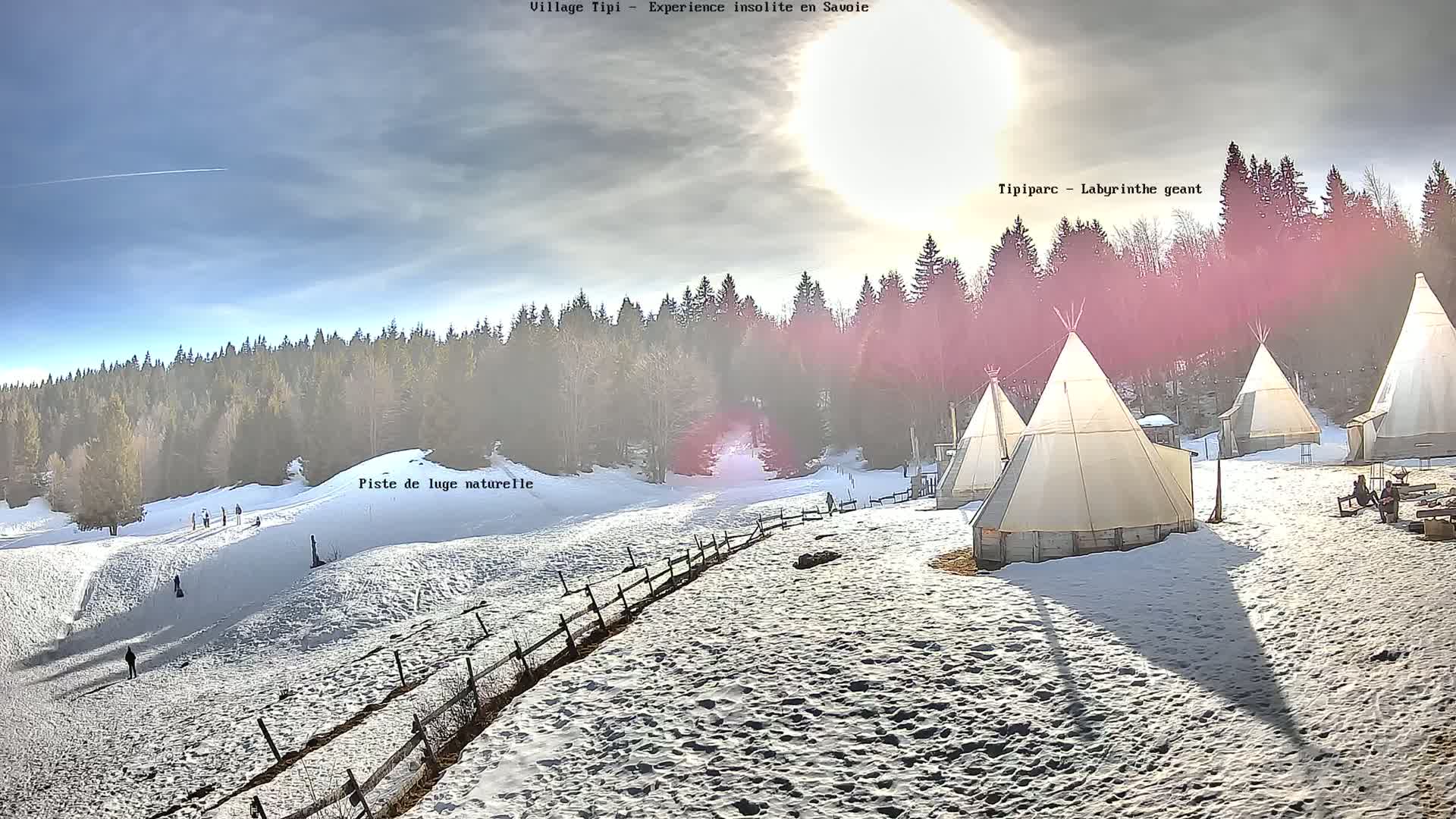 A snowy landscape with several teepees, a wooden fence, and people sledding down a hill under a partly sunny sky.