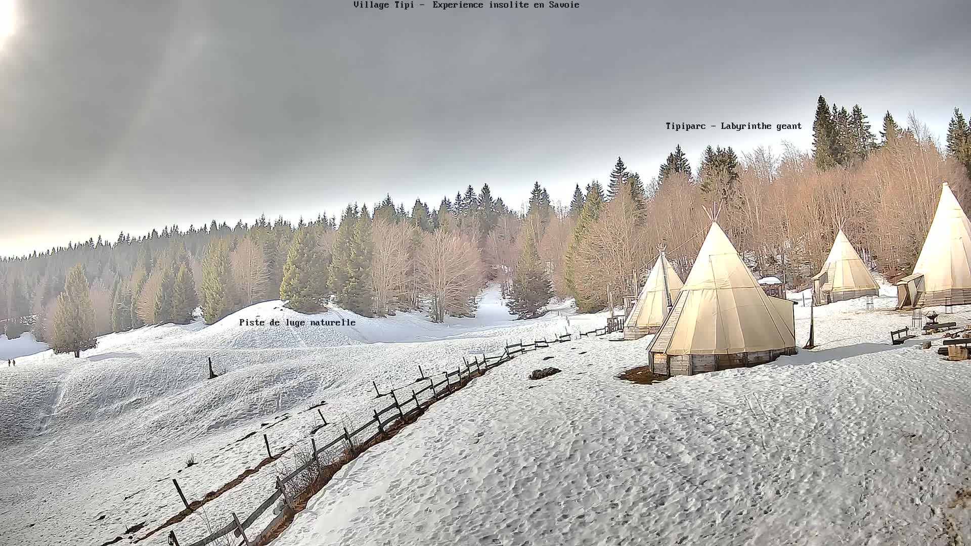 Several teepees are situated on a snow-covered hillside overlooking a forest under a mostly cloudy sky.