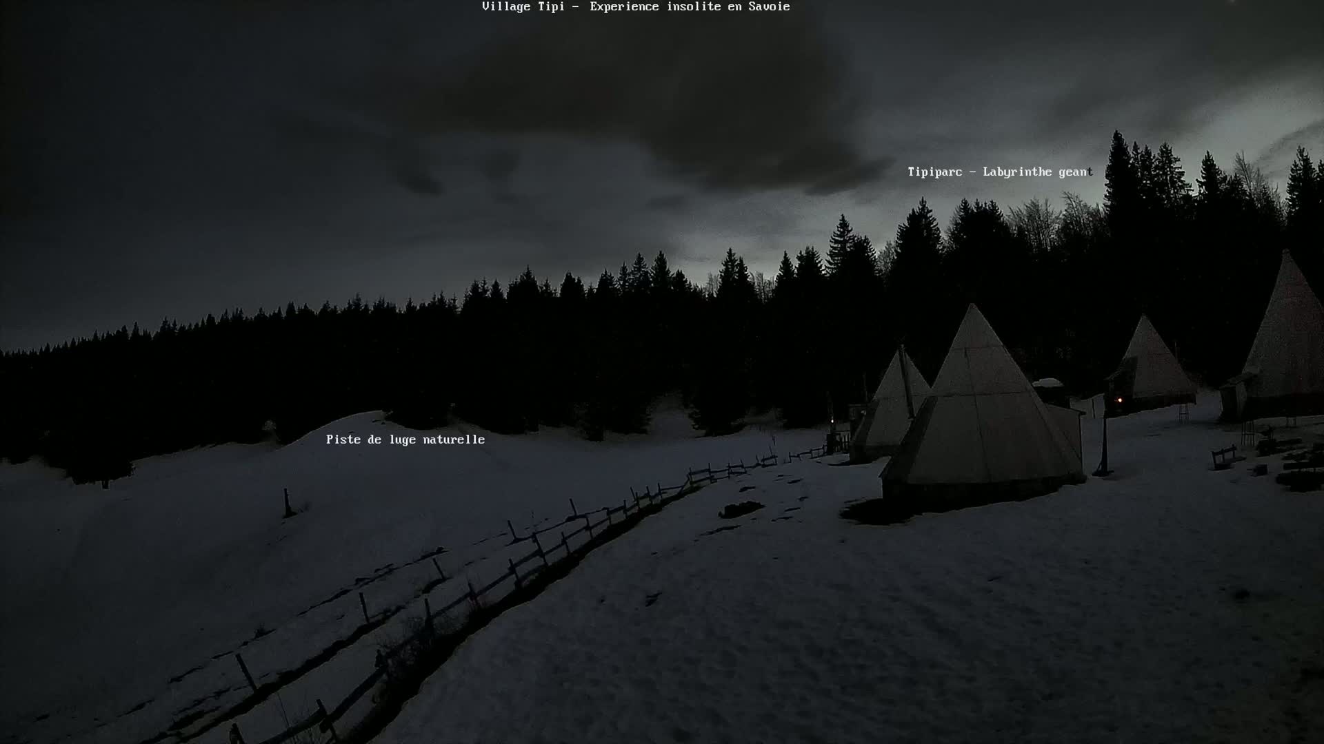 Several teepees are nestled in a snow-covered landscape beneath a dark, cloudy sky at night, surrounded by a dark forest.