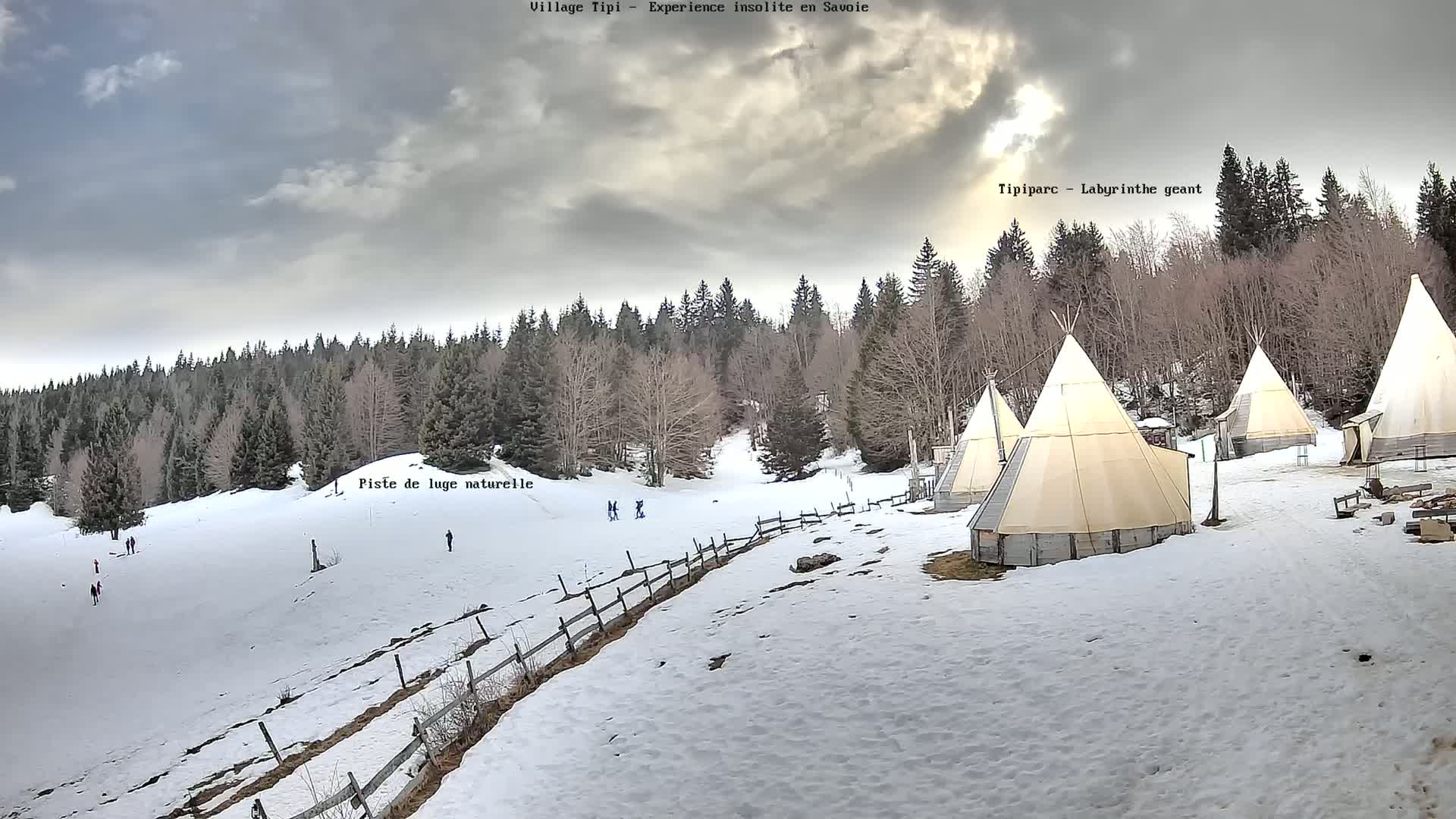 A snow-covered landscape features several teepees nestled among a forest, with a snow-covered toboggan run in the foreground under a cloudy sky.