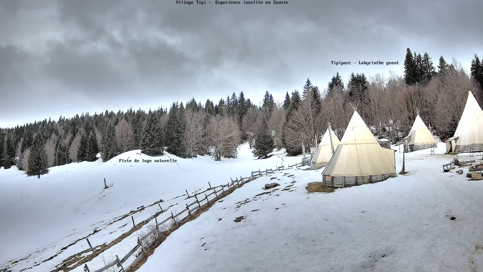 Several teepees are situated on a snow-covered hillside beneath a cloudy sky, with a wooden fence and a forest in the background.