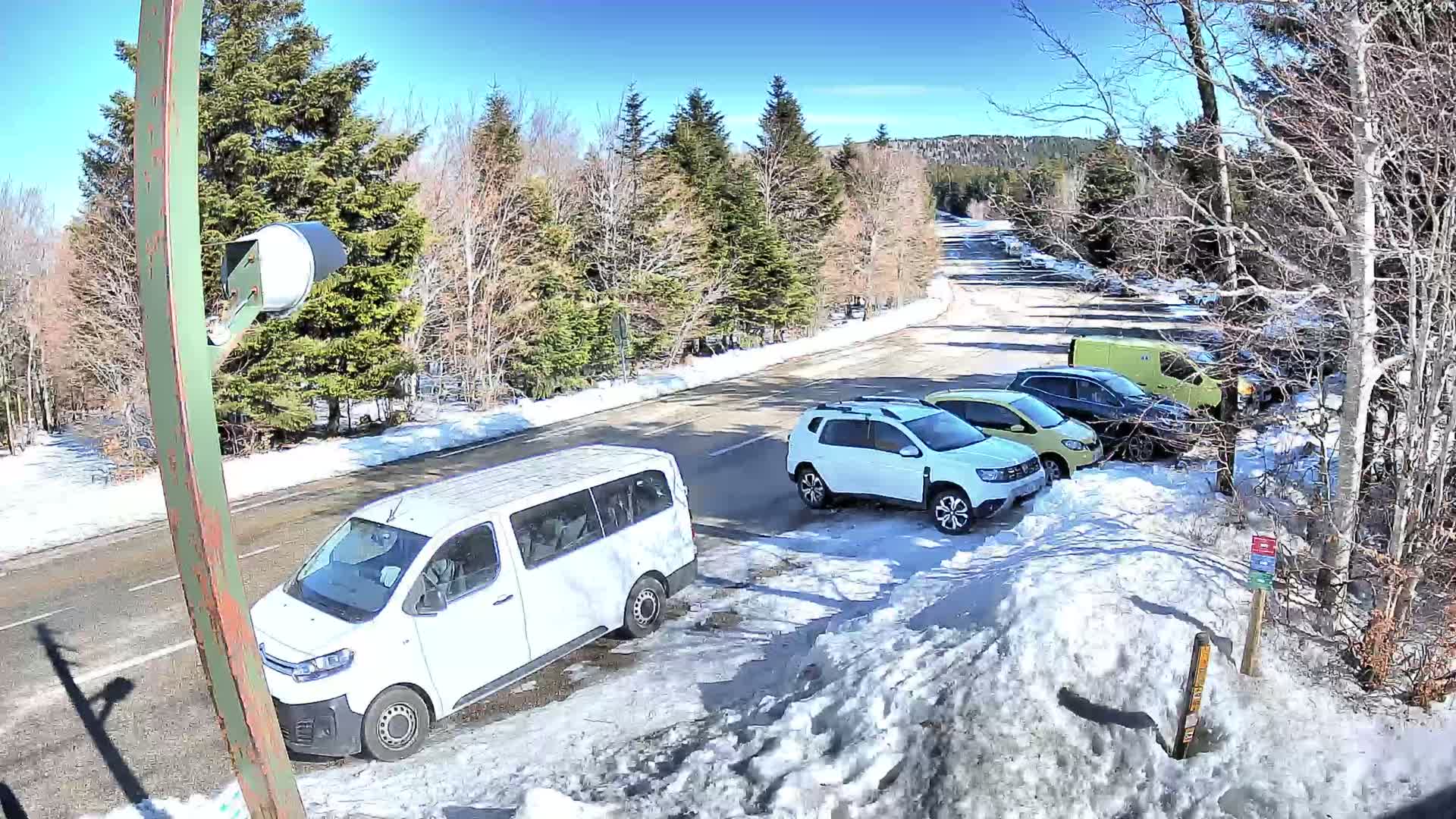A sunny day with patches of snow on the ground shows several vehicles parked along a road bordered by evergreens and deciduous trees.