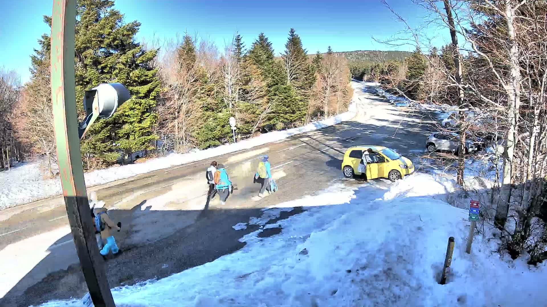 A sunny winter day shows a yellow car parked near a roadway where three people with backpacks cross, while other cars are parked in a lot nearby, surrounded by snow-covered ground and leafless trees.
