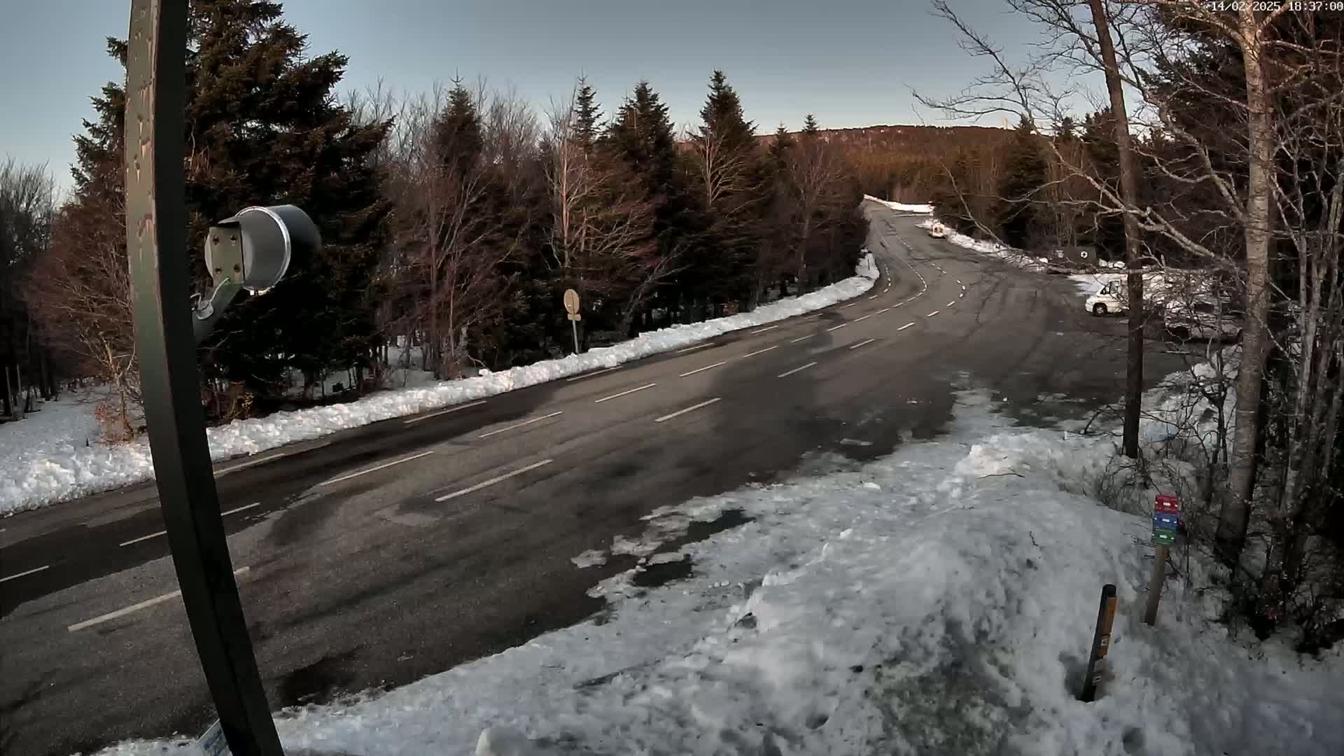 A winding road with snow on the sides curves through a snowy, wooded area under a clear, twilight sky.