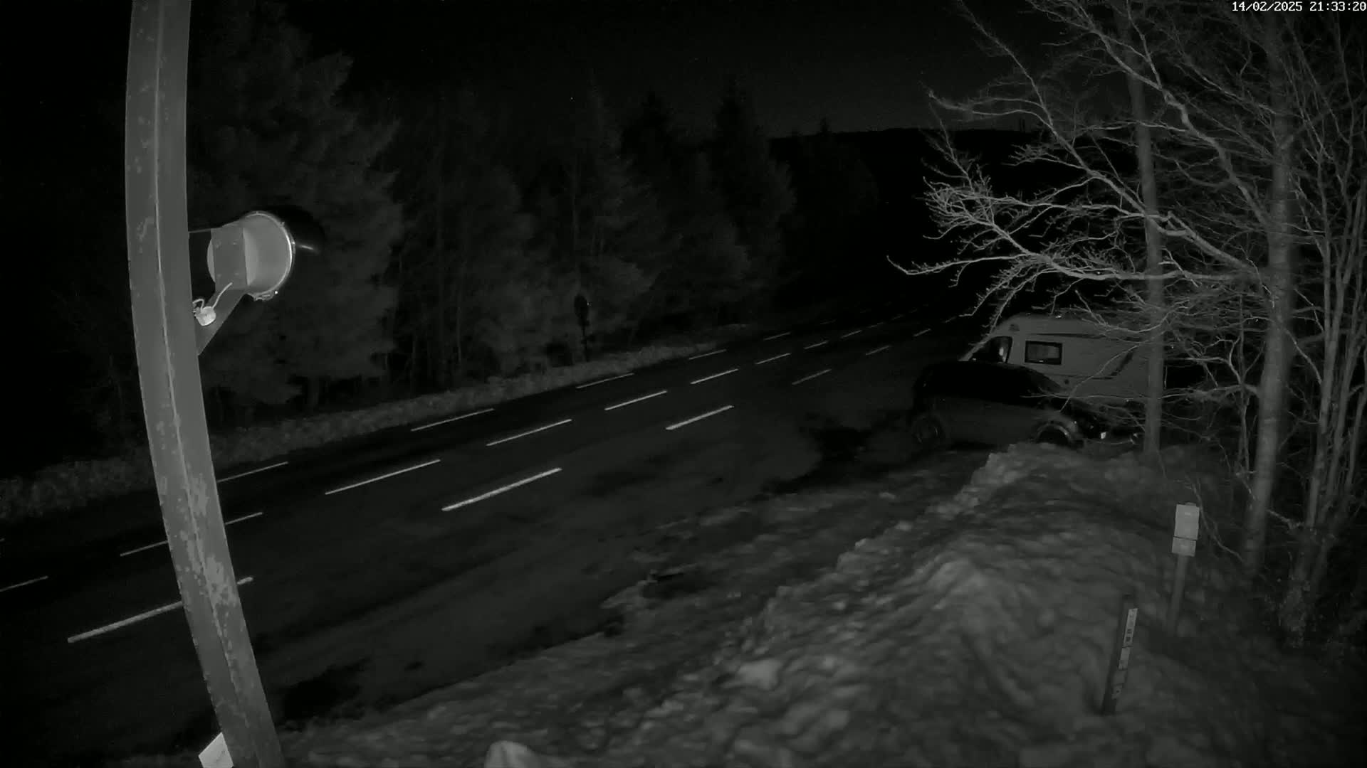 A nighttime, snow-covered roadside scene shows a camper van parked near bare trees beside a dark roadway with white lane markers.