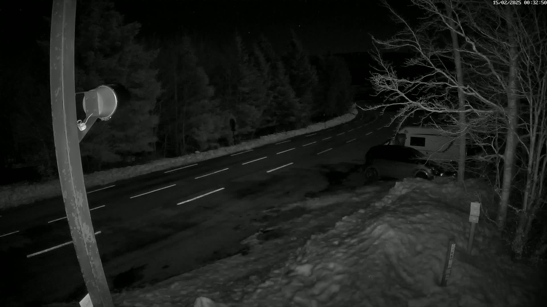 A nighttime, snow-covered road curves through a wooded area, with a car and camper parked nearby.