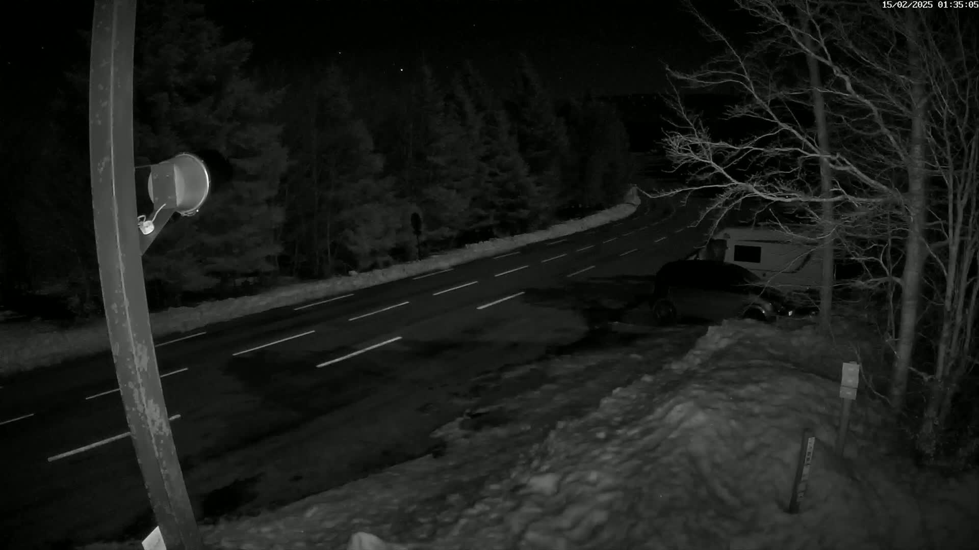 A nighttime view of a snow-covered road curving through a wooded area, with a vehicle parked beside it.