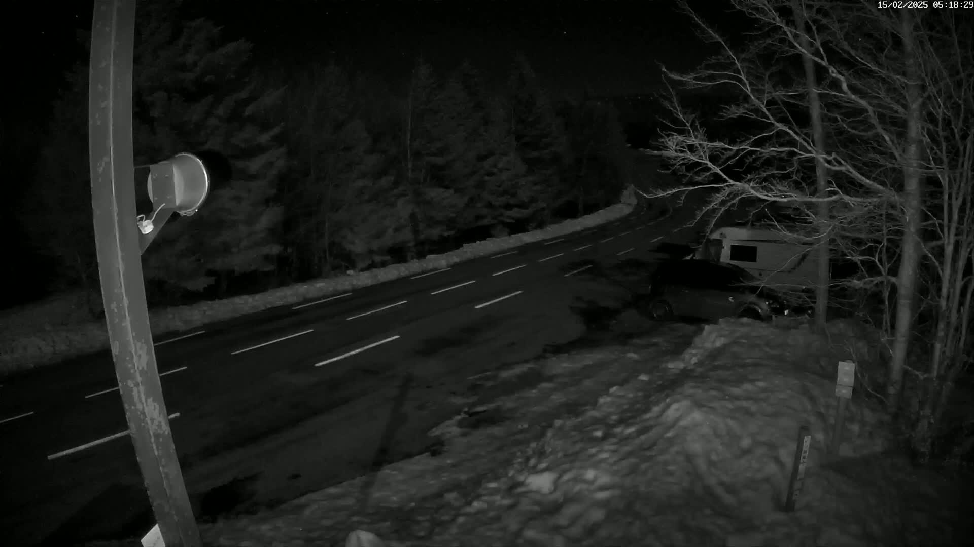 A nighttime, snow-covered roadside scene shows a dark-colored vehicle and a camper parked near a two-lane road lined with trees.