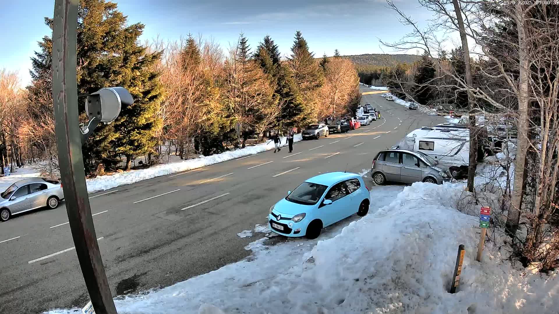 A light-blue car is parked on a snow-covered road next to a parking lot with several other cars and campers, under a mostly sunny sky.