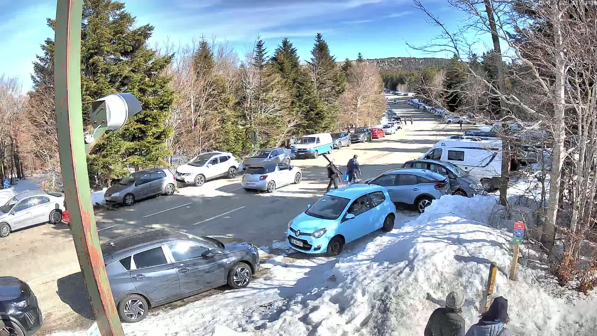 A sunny day shows a crowded parking lot filled with cars in a snow-covered area surrounded by trees.