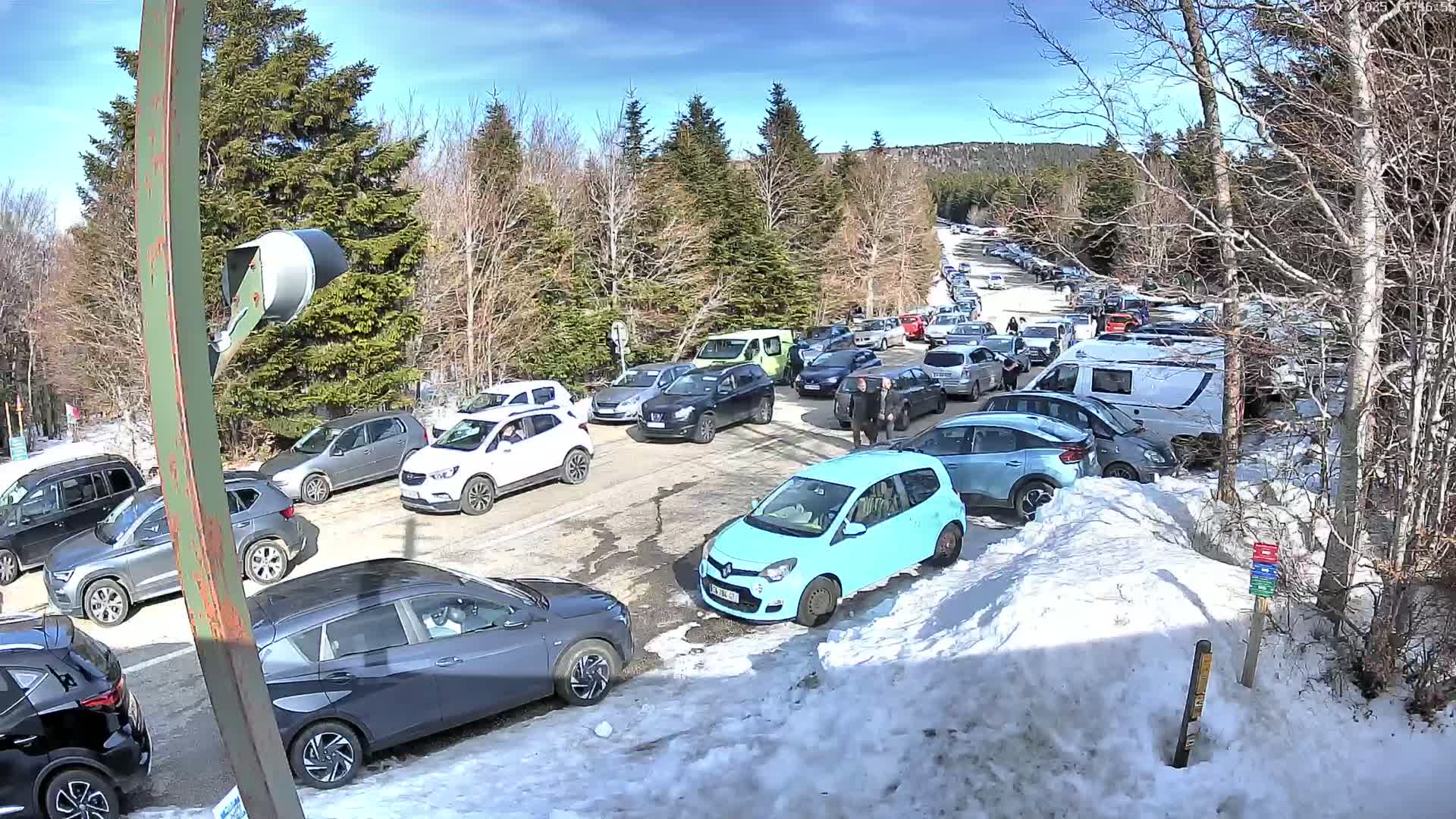 A crowded parking lot with numerous cars parked on a sunny day with snow on the ground and trees in the background.