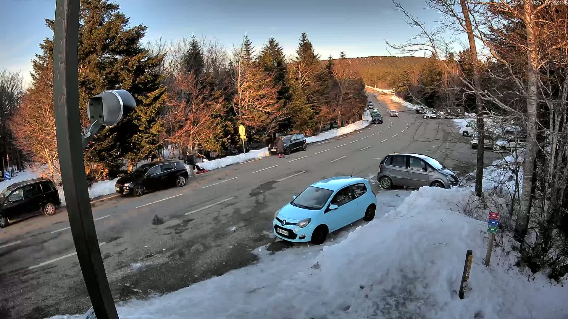 A light-blue car is parked on a snow-covered roadside near several other cars, in a parking area next to a road that winds through a snowy forest under a clear, bright sky.