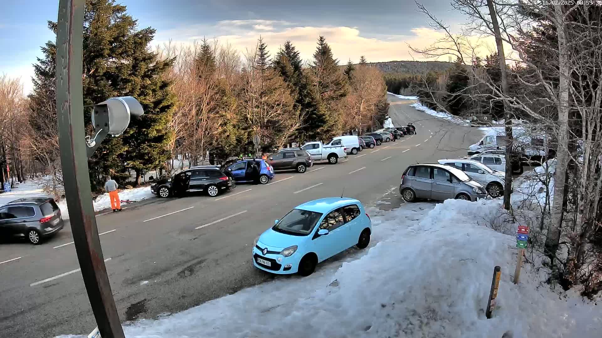 A parking lot with numerous cars parked along a roadway in a snowy, wooded area under a partly cloudy sky.