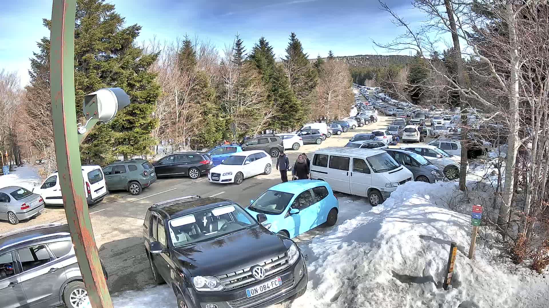 A large parking lot filled with cars, some people, and snow under a sunny sky.