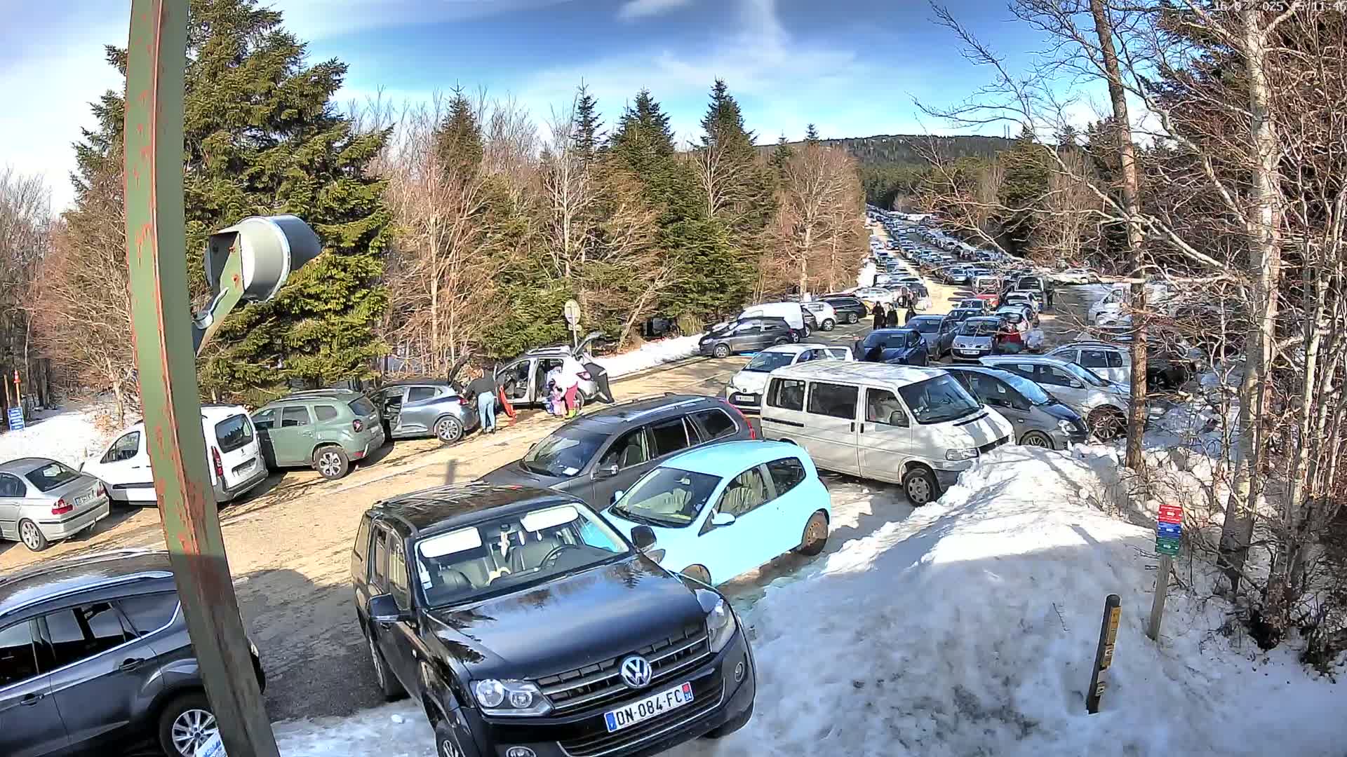 A large parking lot filled with cars is surrounded by snow and bare deciduous trees under a sunny sky.