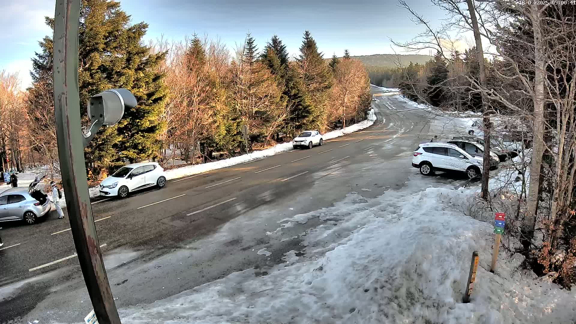 A paved road winds through a snowy, wooded area with several parked cars and a few people near a vehicle.