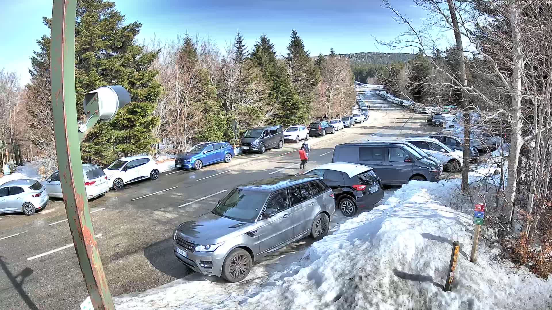 A parking lot filled with cars, bordered by snow banks and evergreen trees, under a sunny sky.