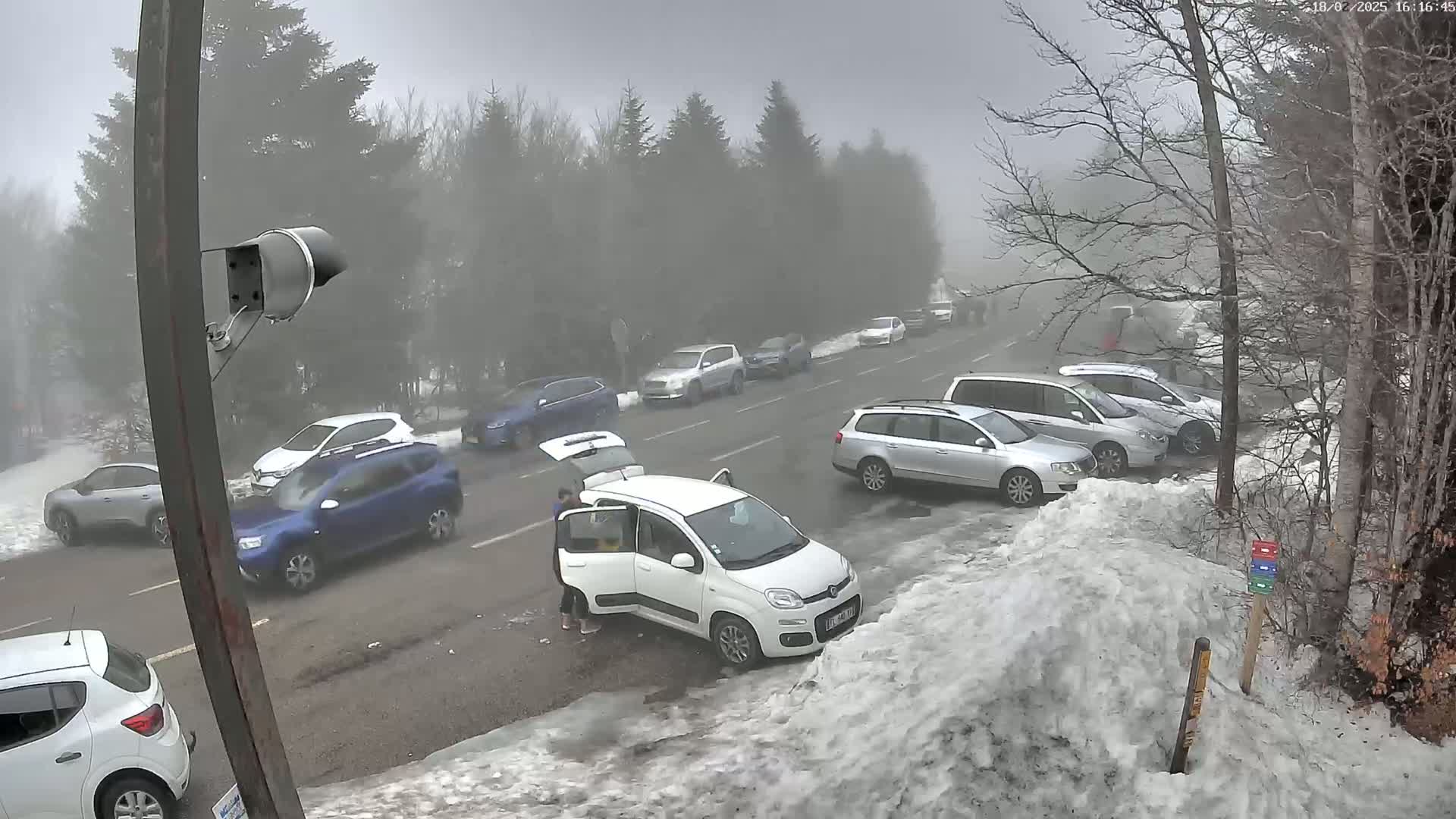 A foggy parking lot with several cars parked in it and some cars driving on a road next to a snow bank.