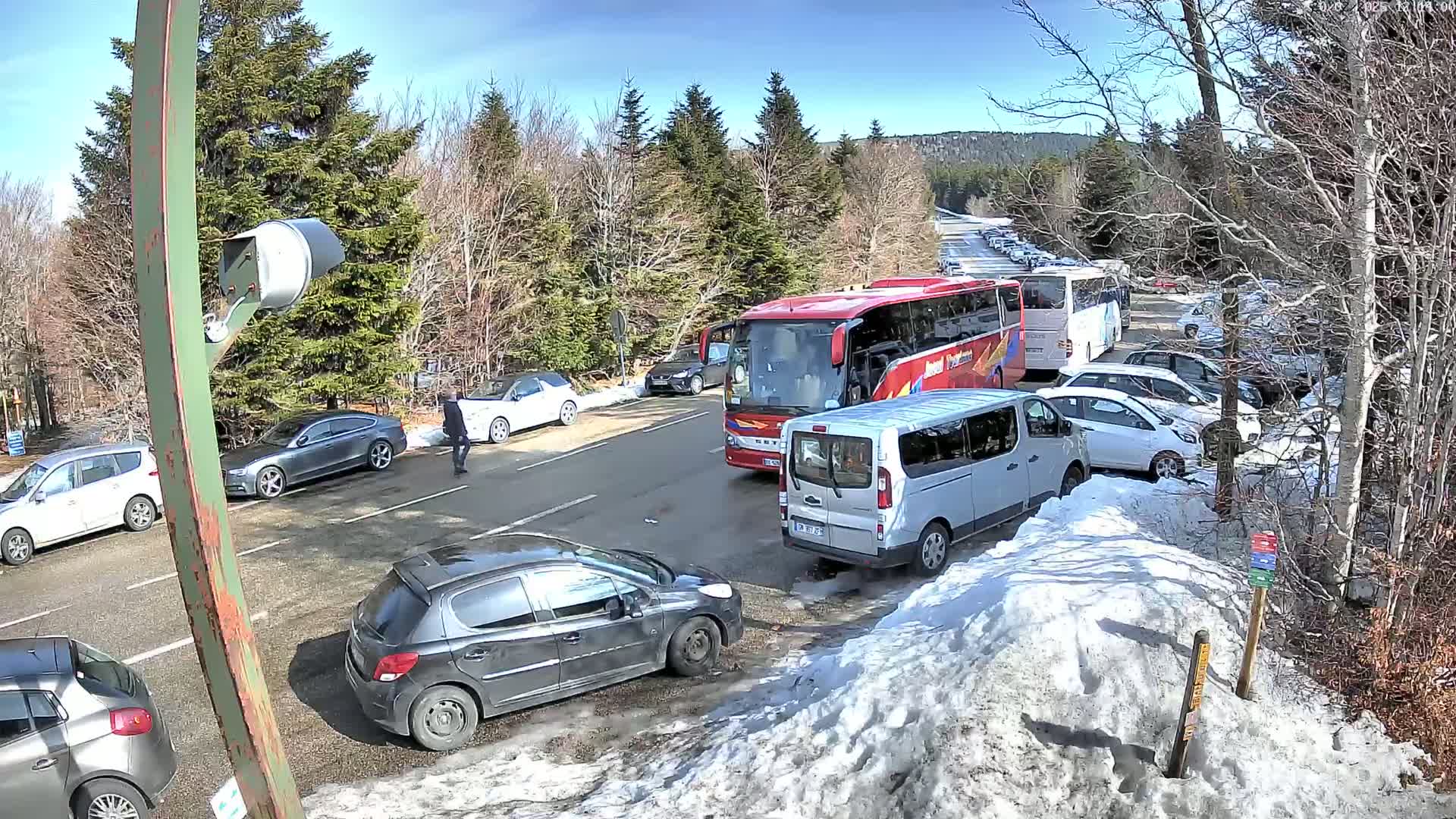 A sunny day shows a parking lot with numerous cars and buses, some snow on the ground, and a wooded area in the background.
