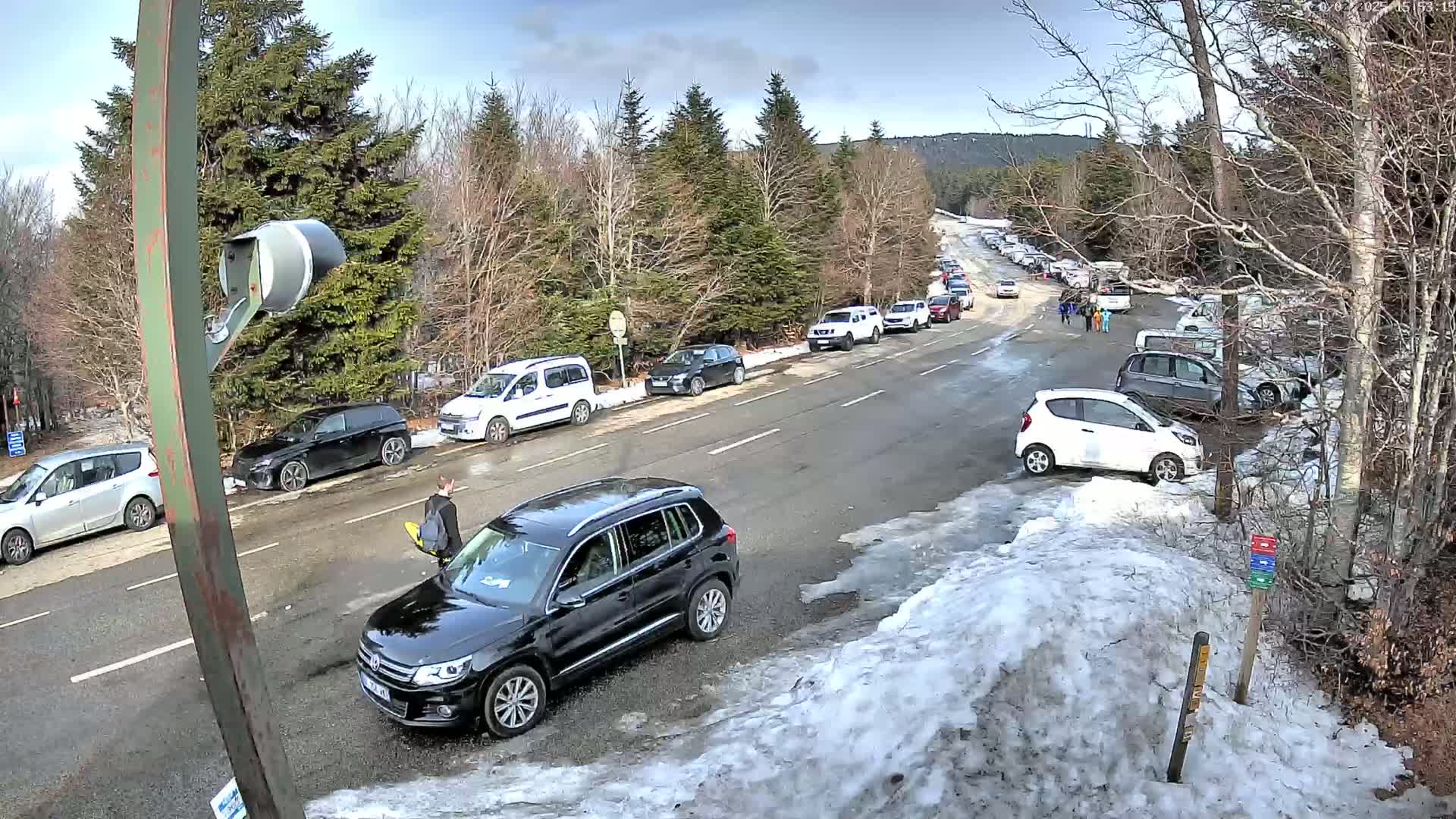 A parking lot and road lined with cars are partially covered in snow under a partly cloudy sky, with a few people walking nearby.