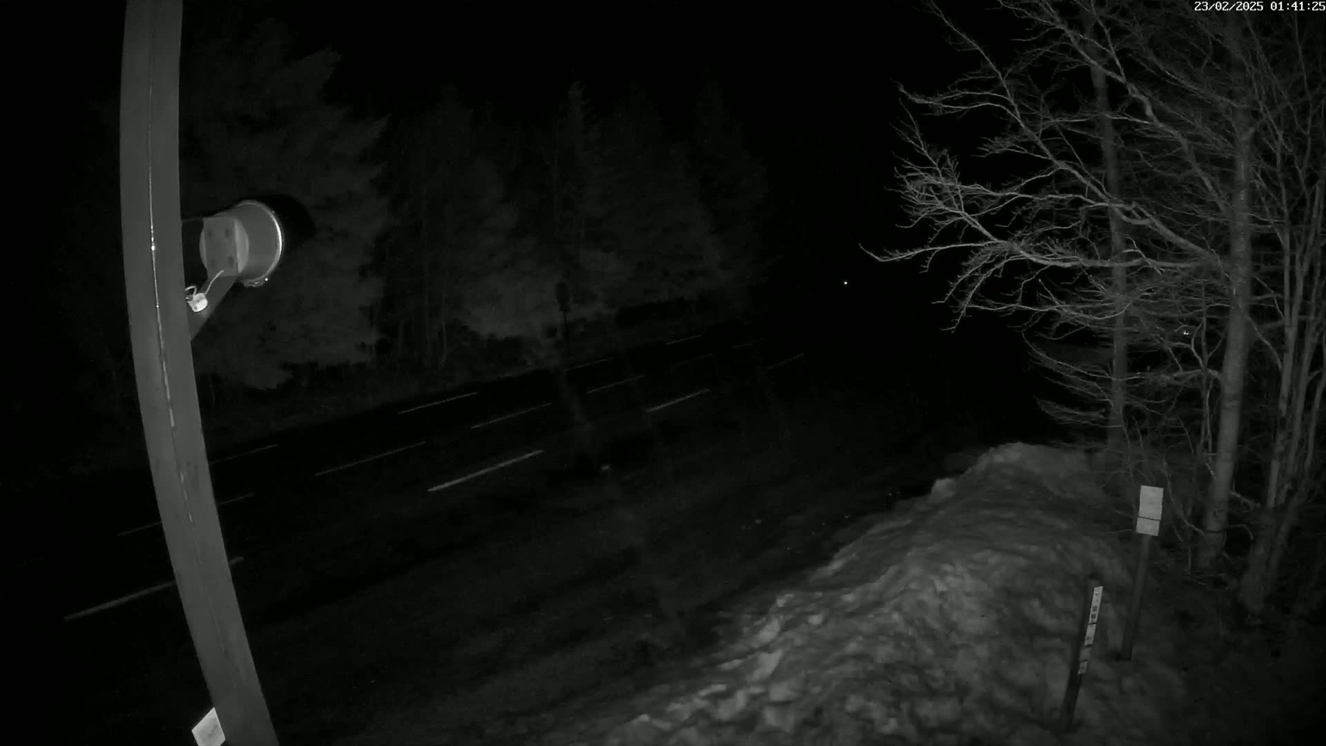 A nighttime, snow-covered roadside scene shows a dark road with white lane markings, bare trees, and a metal post with a device attached.