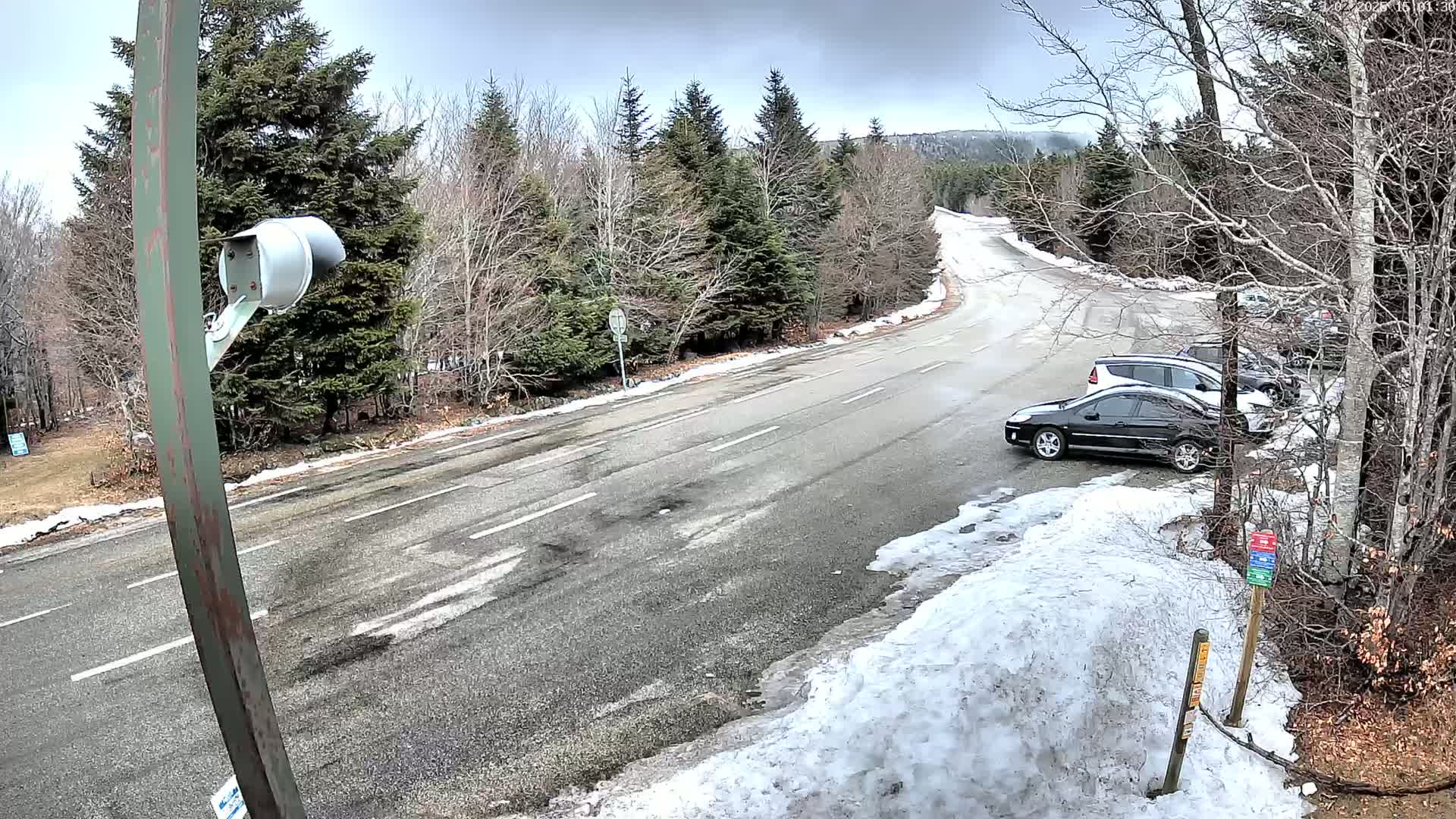 A paved road curves through a snowy, wooded area with several cars parked nearby under an overcast sky.