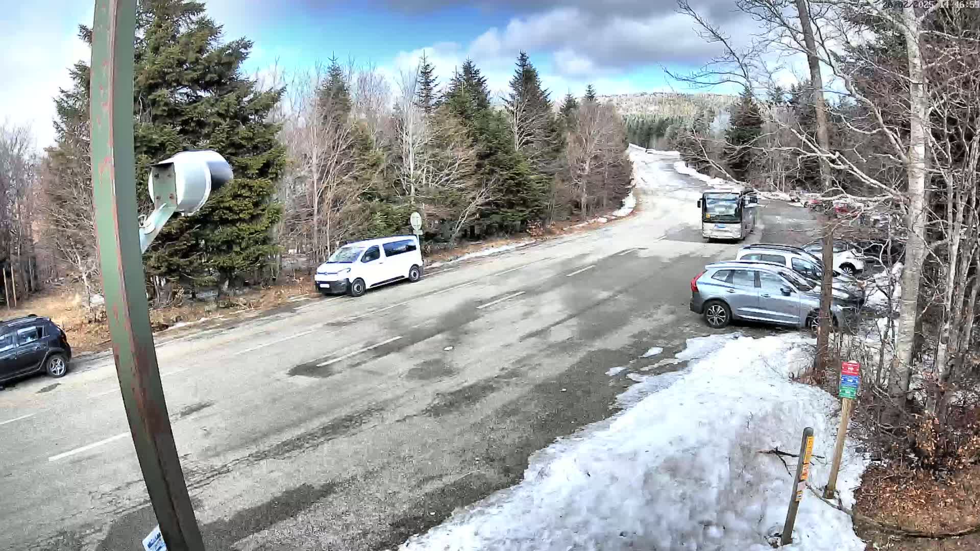 A partly cloudy day shows a roadway with several vehicles parked and driving, some snow on the ground, and a background of bare deciduous trees and evergreens.