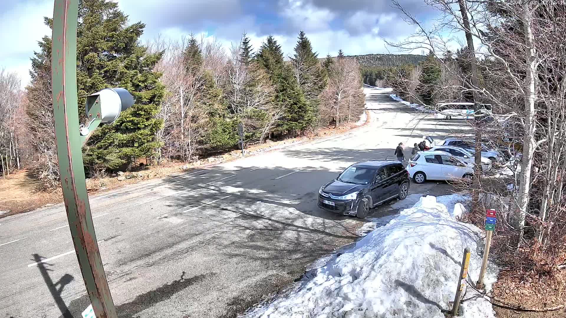 A sunny day with patchy snow shows a road curving through a parking lot with several cars and people, surrounded by leafless trees and evergreens.