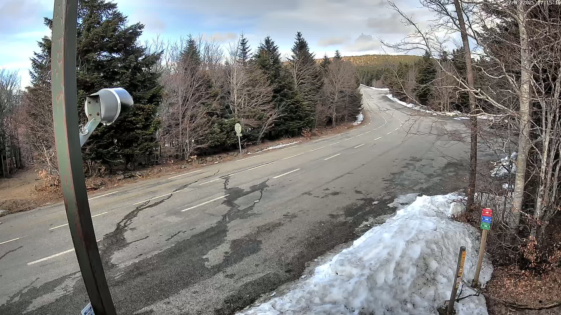 A mostly cloudy day reveals a paved road winding through a forest with snow banks at the edges, under a partly cloudy sky.