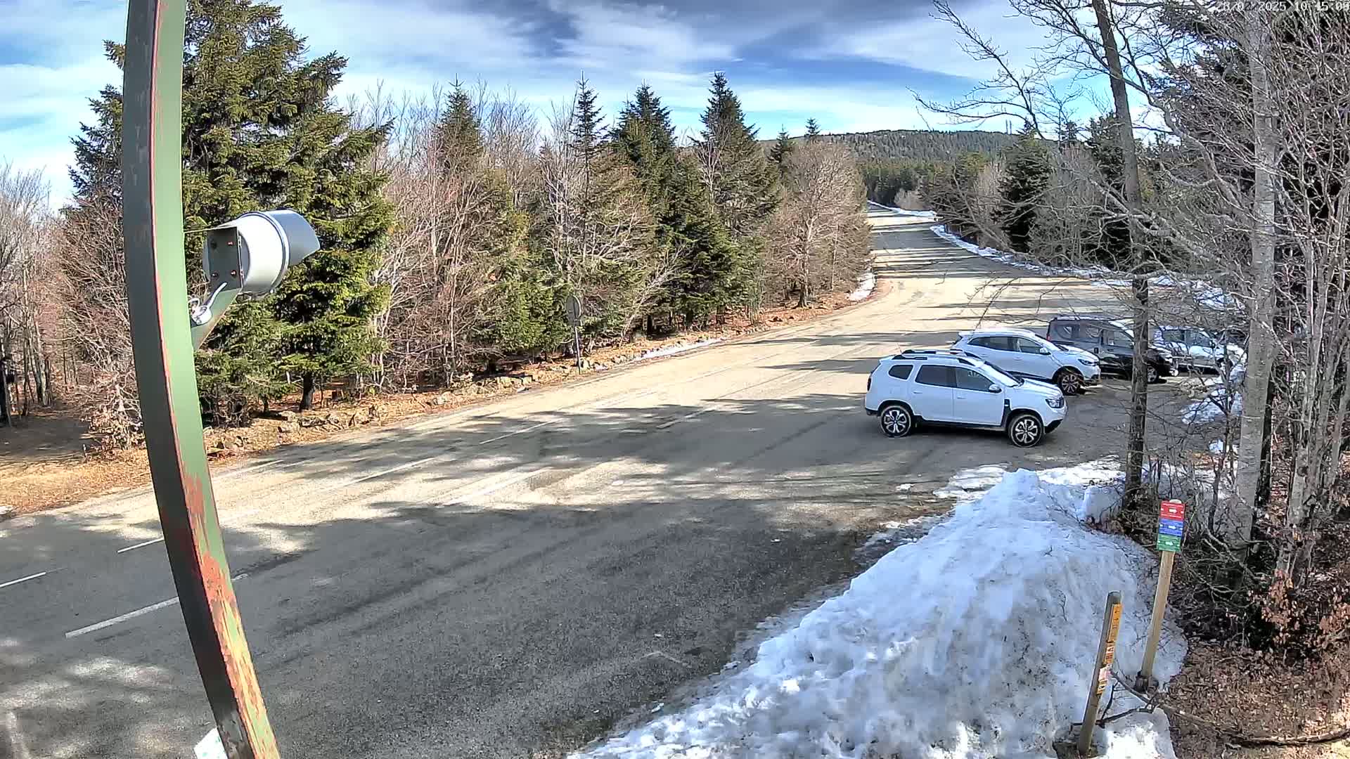 A paved road winds through a snow-dusted forest on a partly sunny day, with several cars parked along its side.