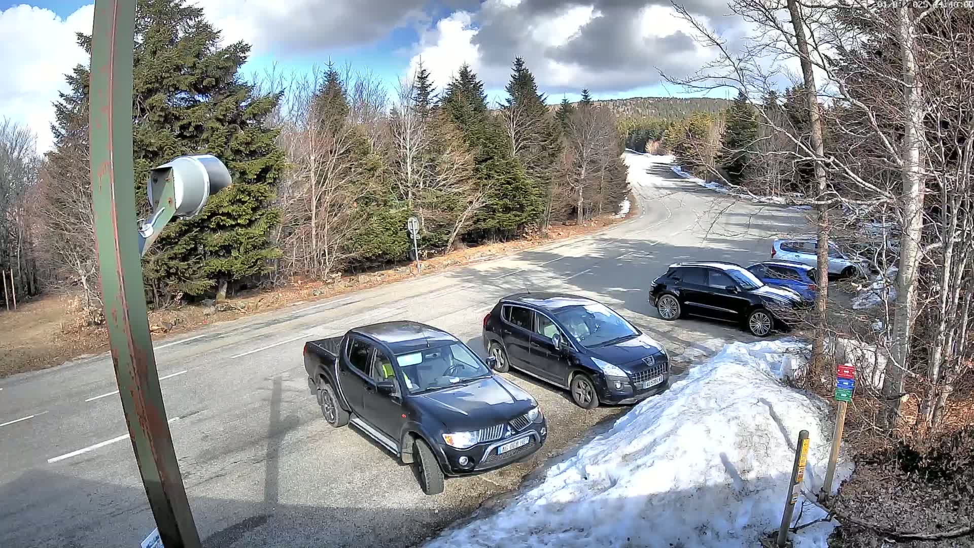 A parking lot with several dark-colored vehicles is situated near a road winding through a sparsely wooded area on a partly sunny day with some snow on the ground.
