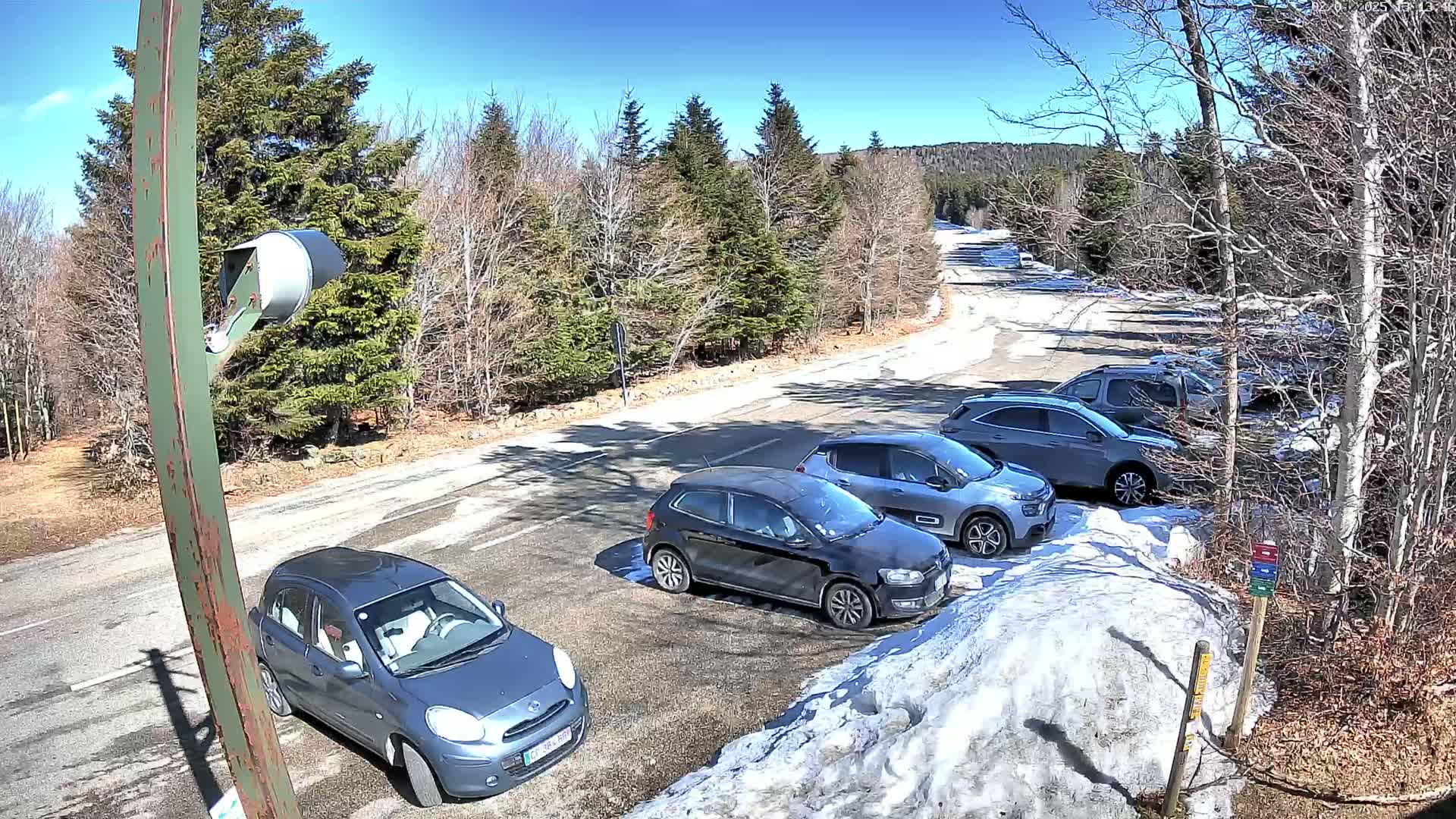 A sunny day in a parking lot surrounded by snow and evergreen trees, with several cars parked.